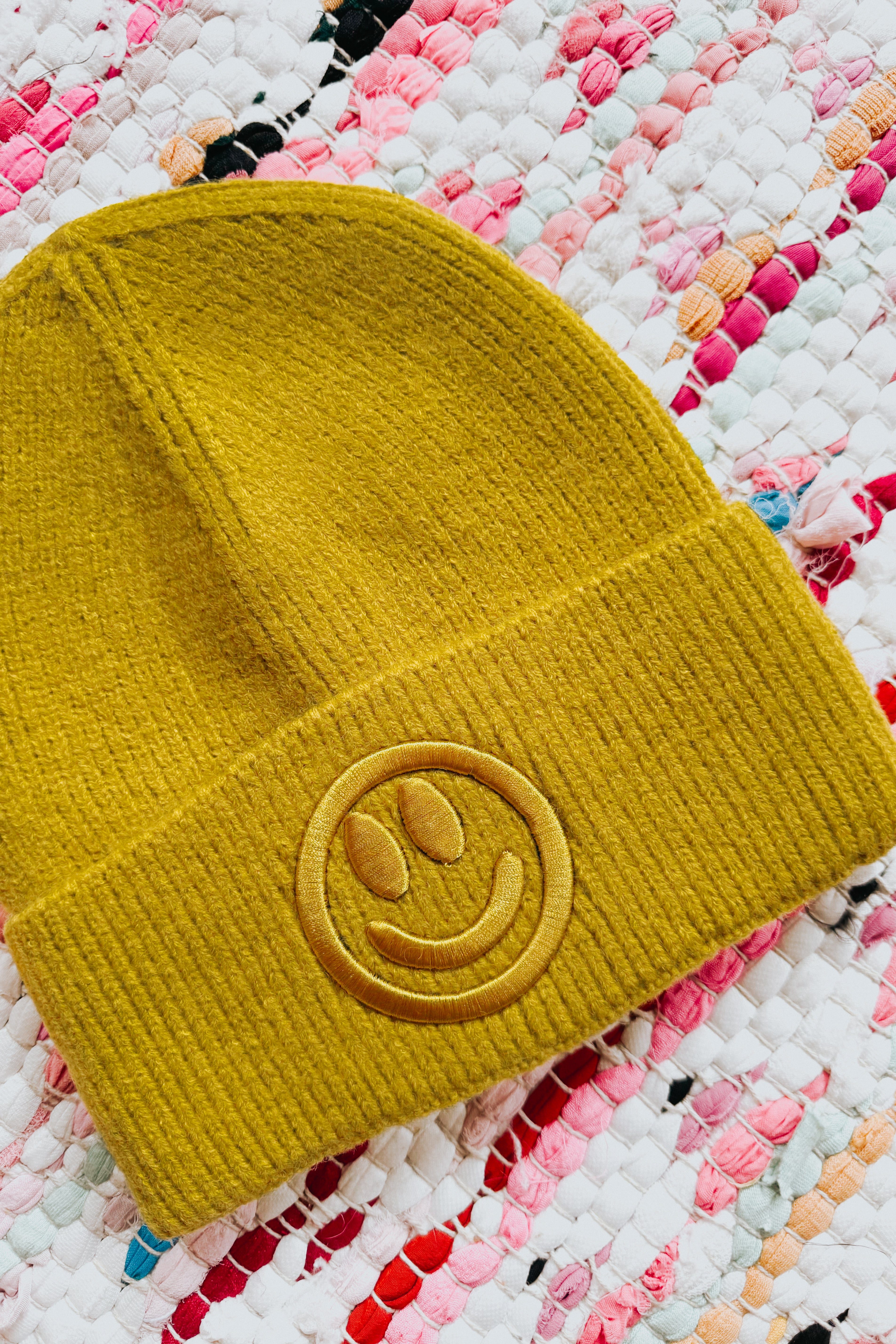 Mustard yellow ribbed knit beanie with embroidered smiley face resting on colorful woven rug background