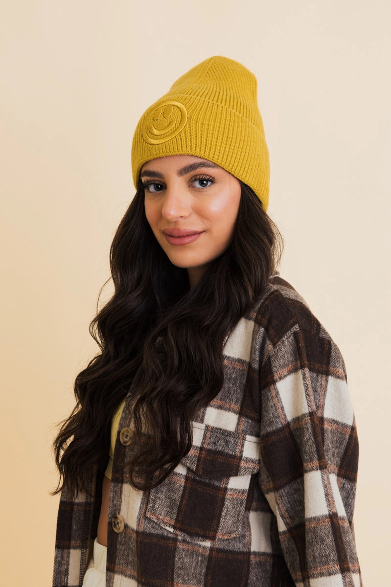 Woman wearing mustard yellow ribbed knit beanie with raised smiley face patch and brown plaid jacket against beige background