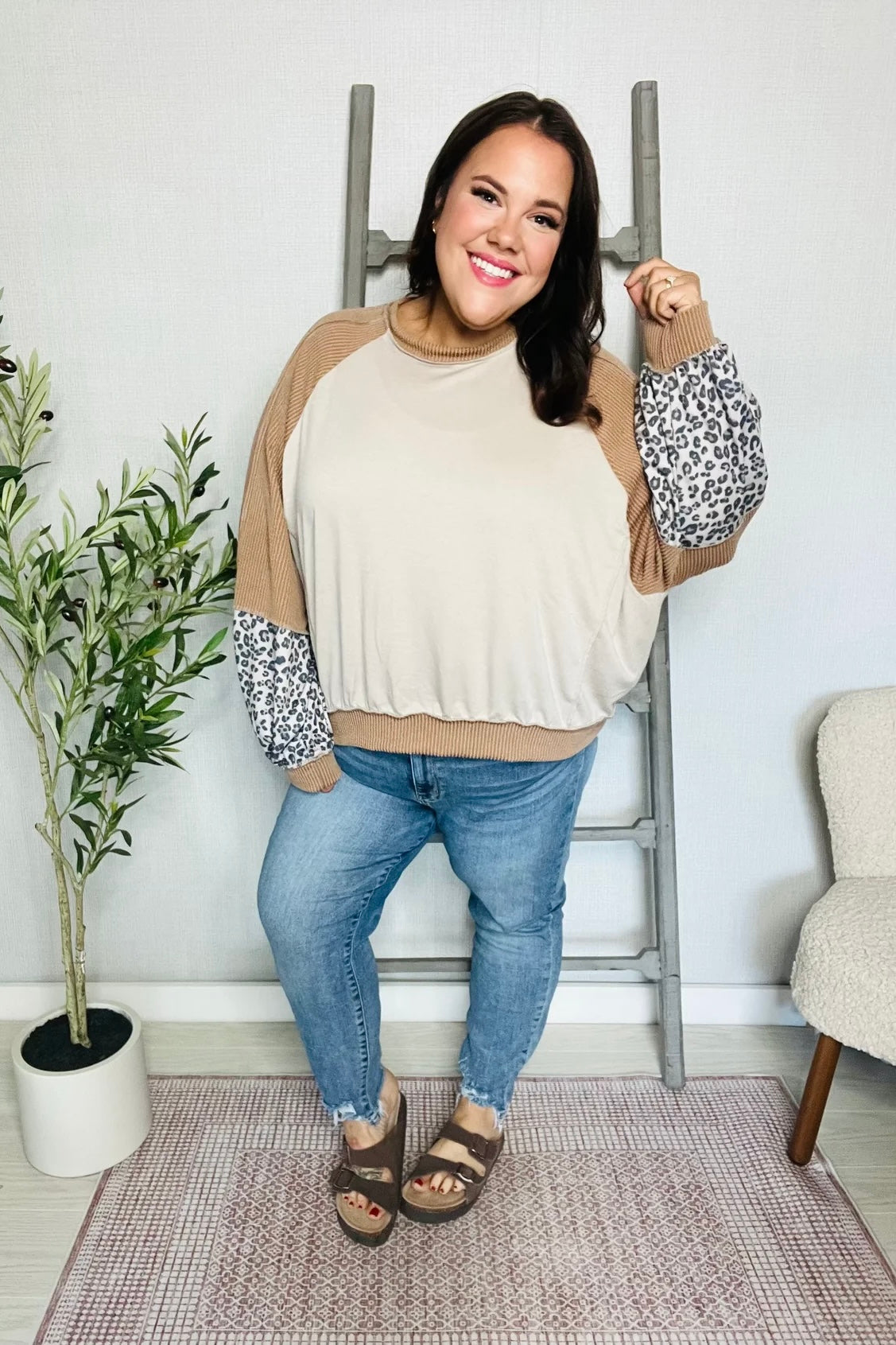 Smiling woman posing in beige and camel dolman colorblock top with leopard print sleeves and blue jeans indoors