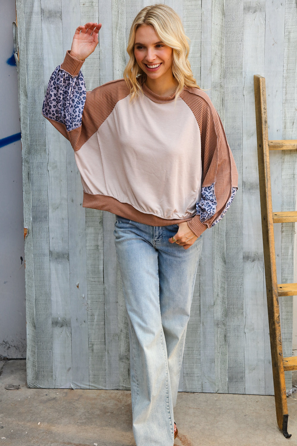 Blonde woman wearing beige and brown colorblock dolman top with leopard print puff sleeves and light wash jeans standing by wooden wall
