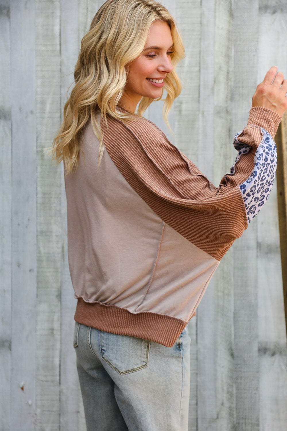 Blonde woman wearing beige and brown dolman colorblock top with ribbed and leopard print sleeve details by wooden fence