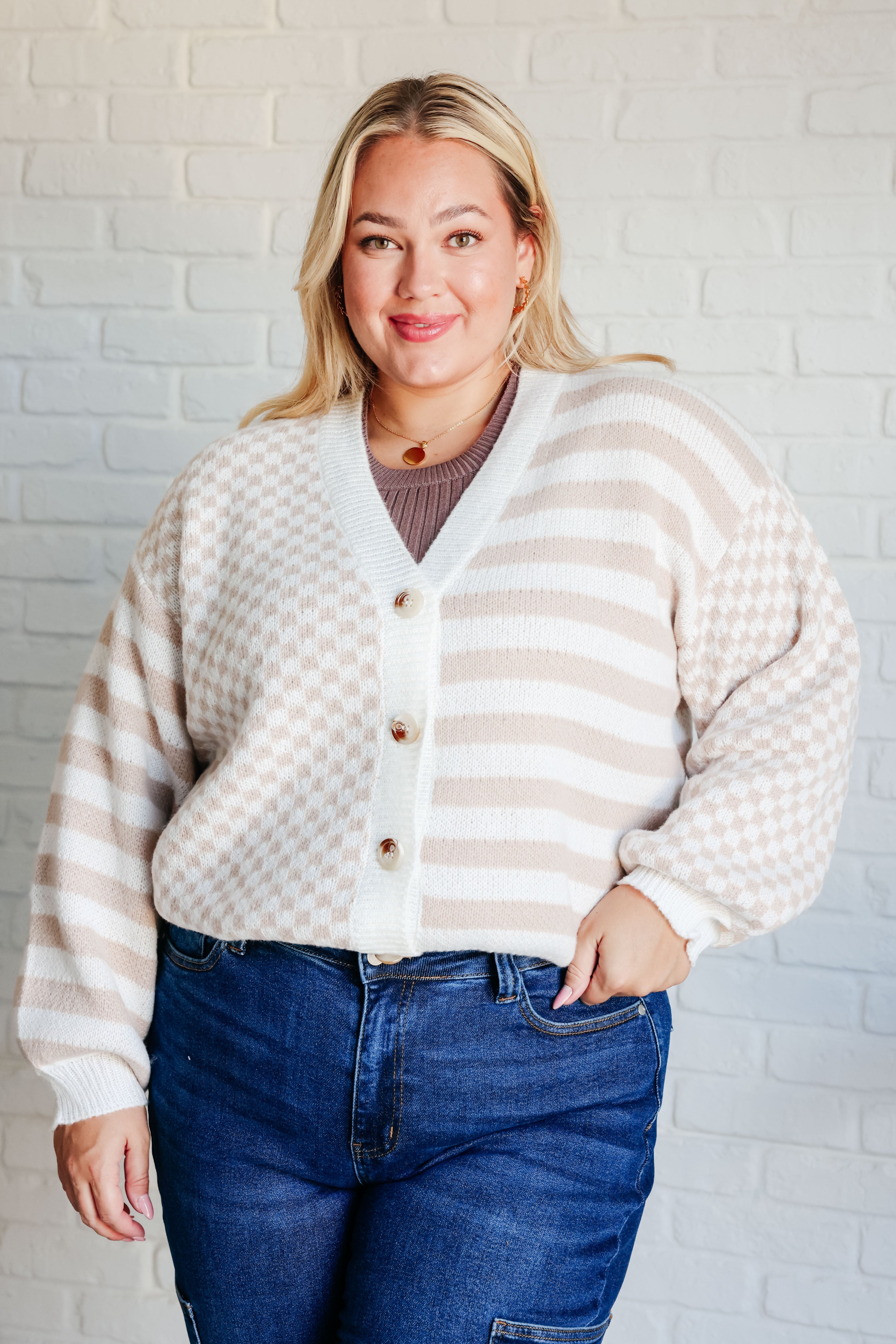 Woman wearing beige and white cardigan with mixed checkered and striped pattern and blue jeans against white brick wall