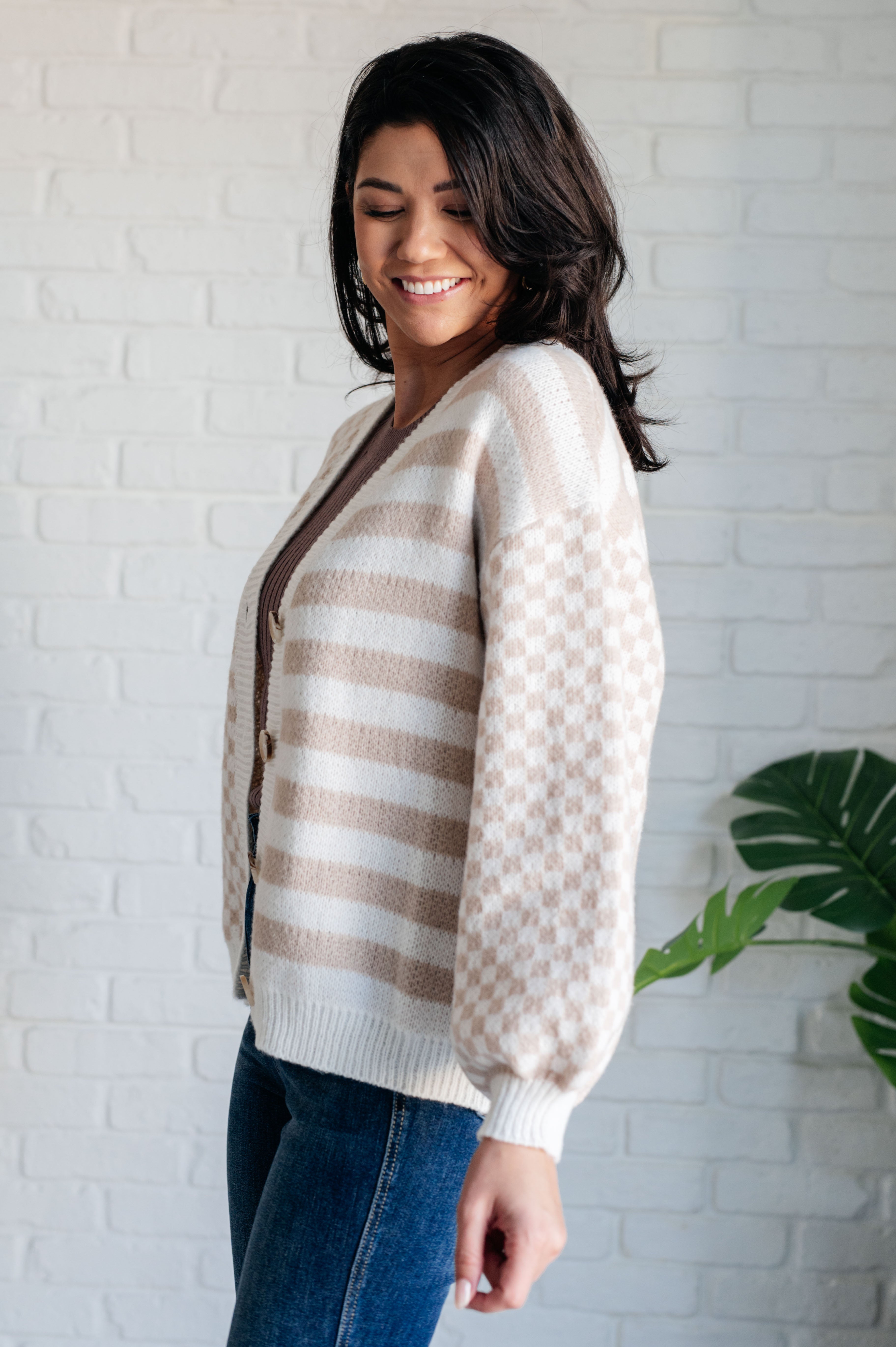 Woman wearing beige and white striped cardigan with checkered puff sleeves and blue jeans standing indoors against white brick wall