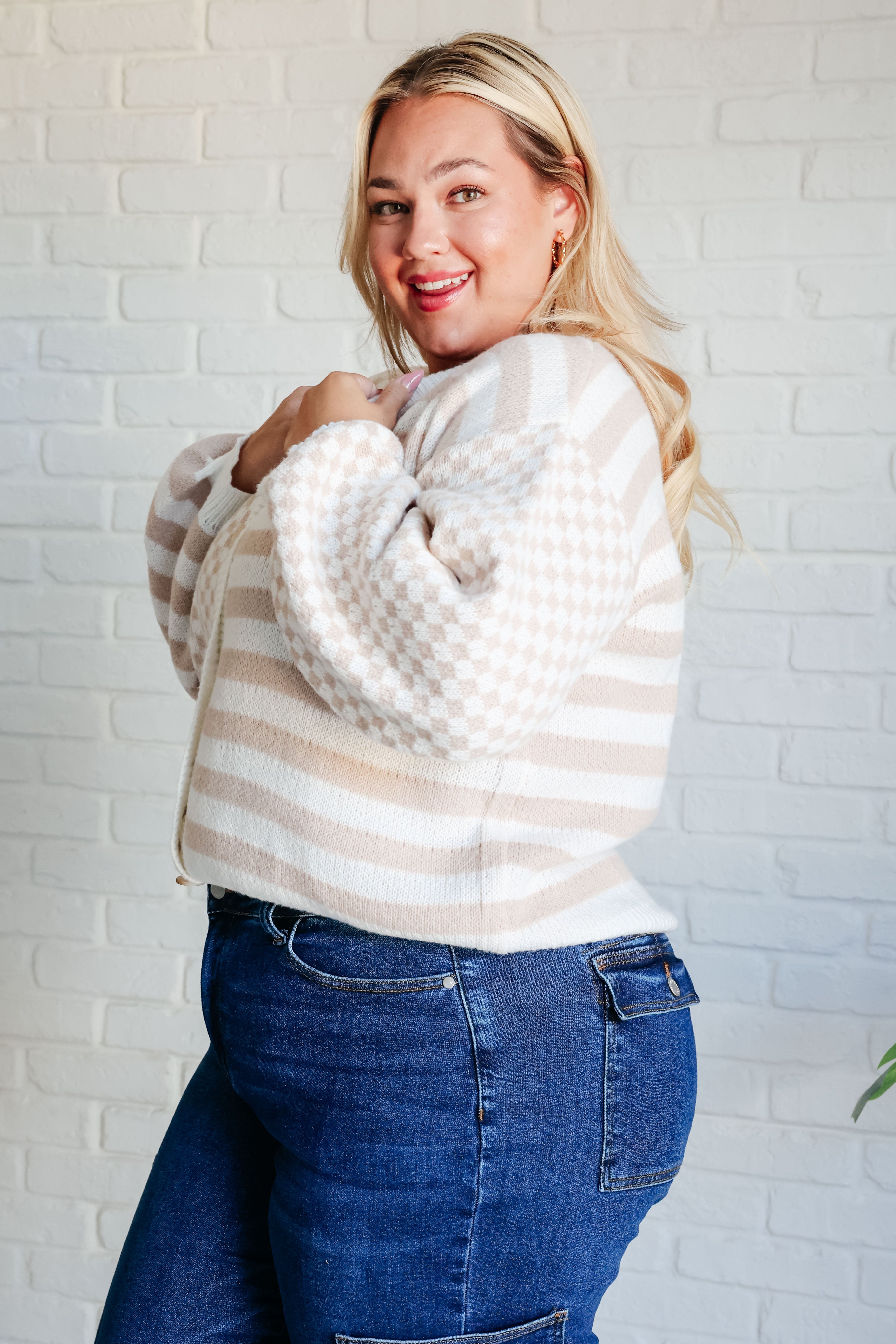 Smiling woman wearing beige and white striped cardigan with checkered balloon sleeves and dark blue jeans standing indoors