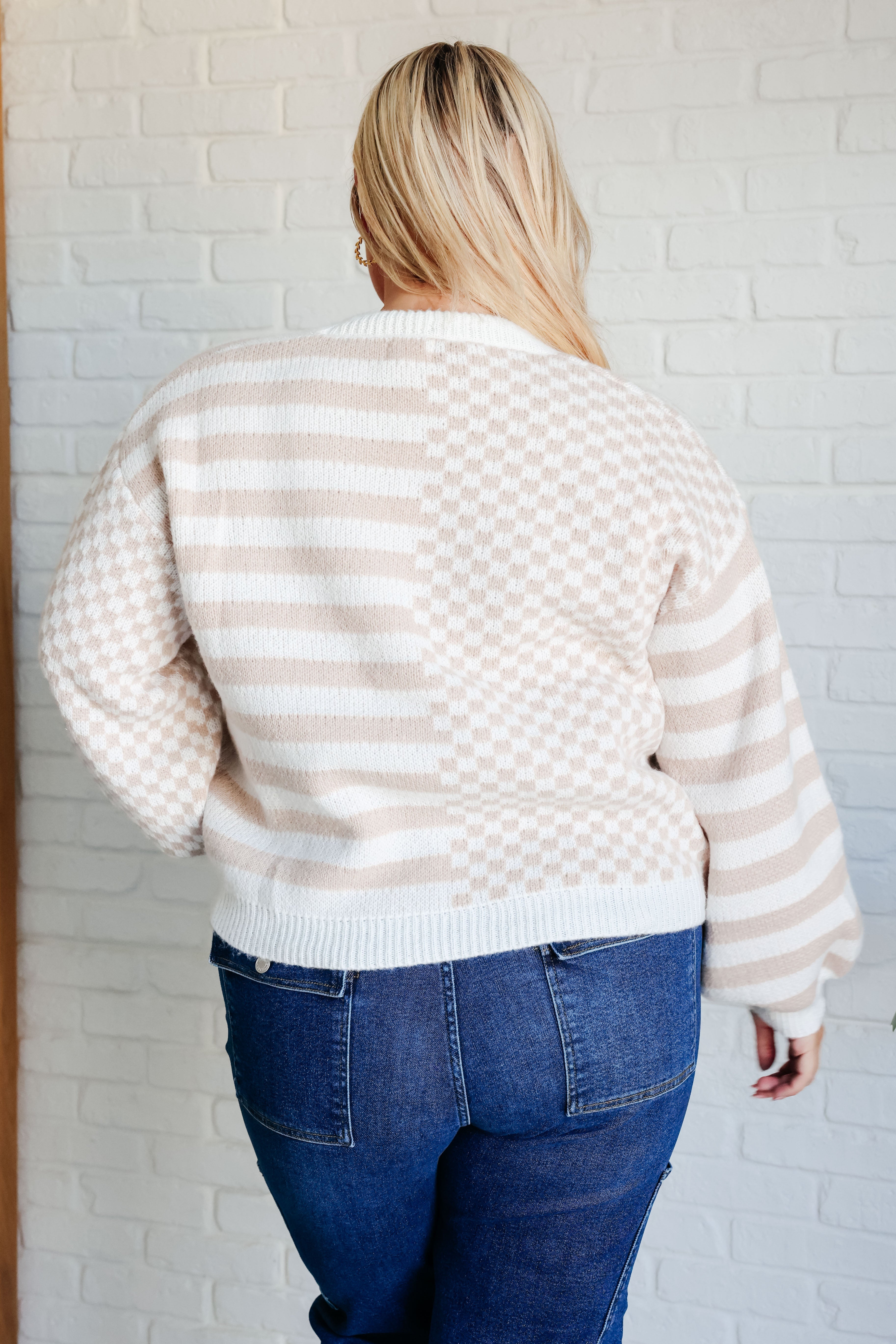Woman wearing beige and white checkered and striped cardigan with balloon sleeves and dark blue jeans standing against white brick wall