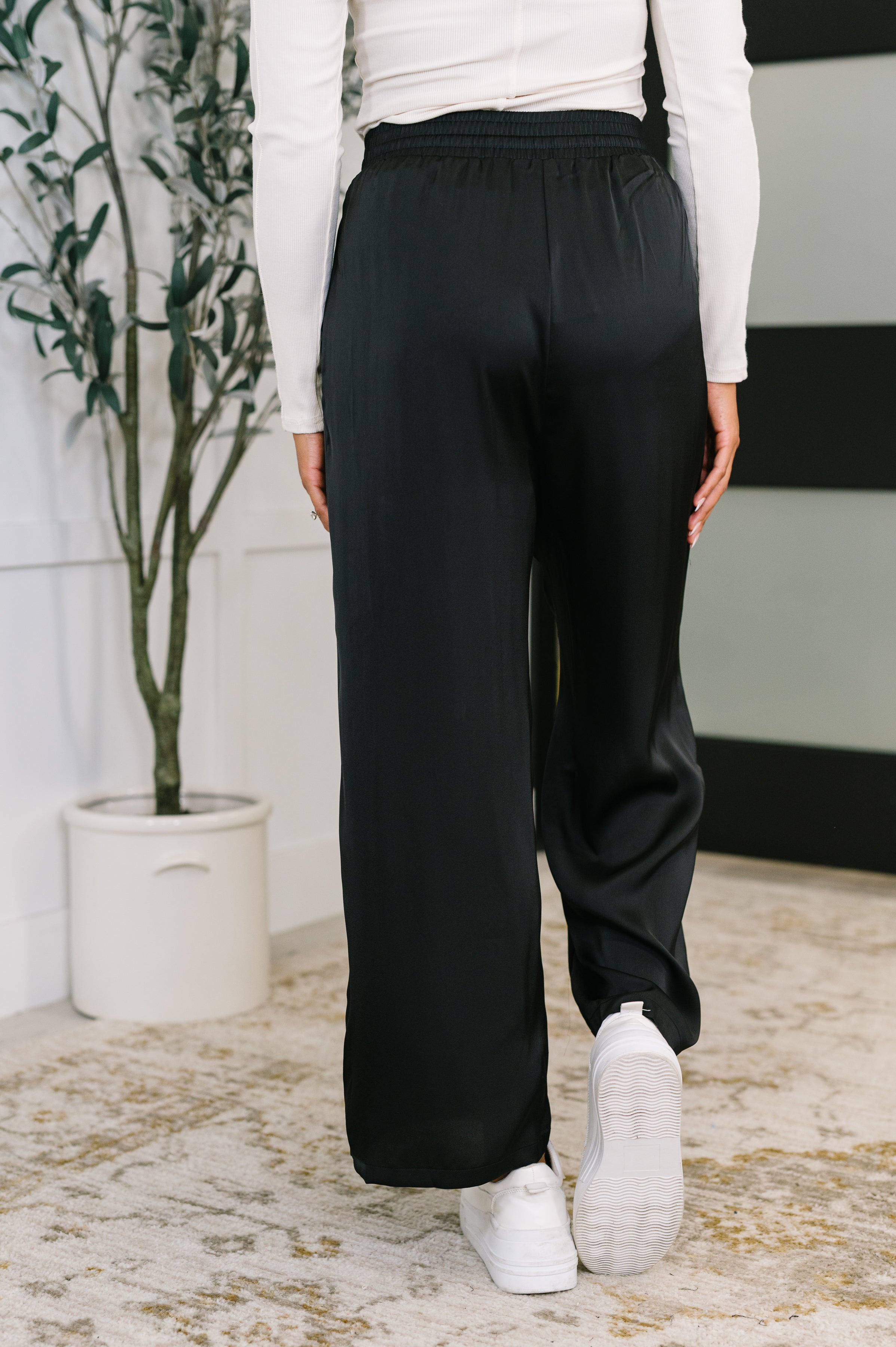 Rear view of person wearing black wide leg pants paired with white long sleeve top and white sneakers indoors