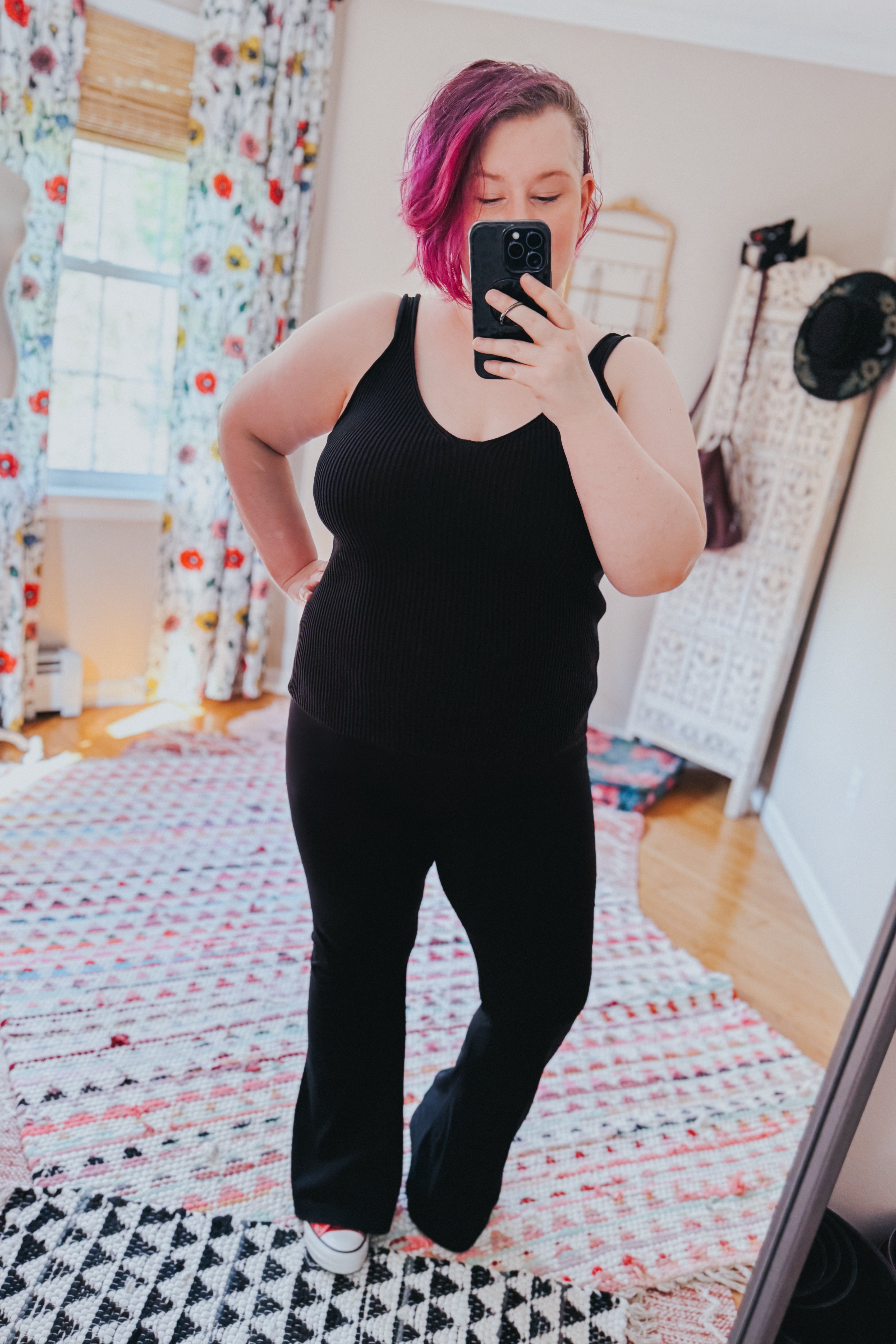Woman with pink short hair wearing black ribbed tank top and black pants taking selfie in bedroom mirror