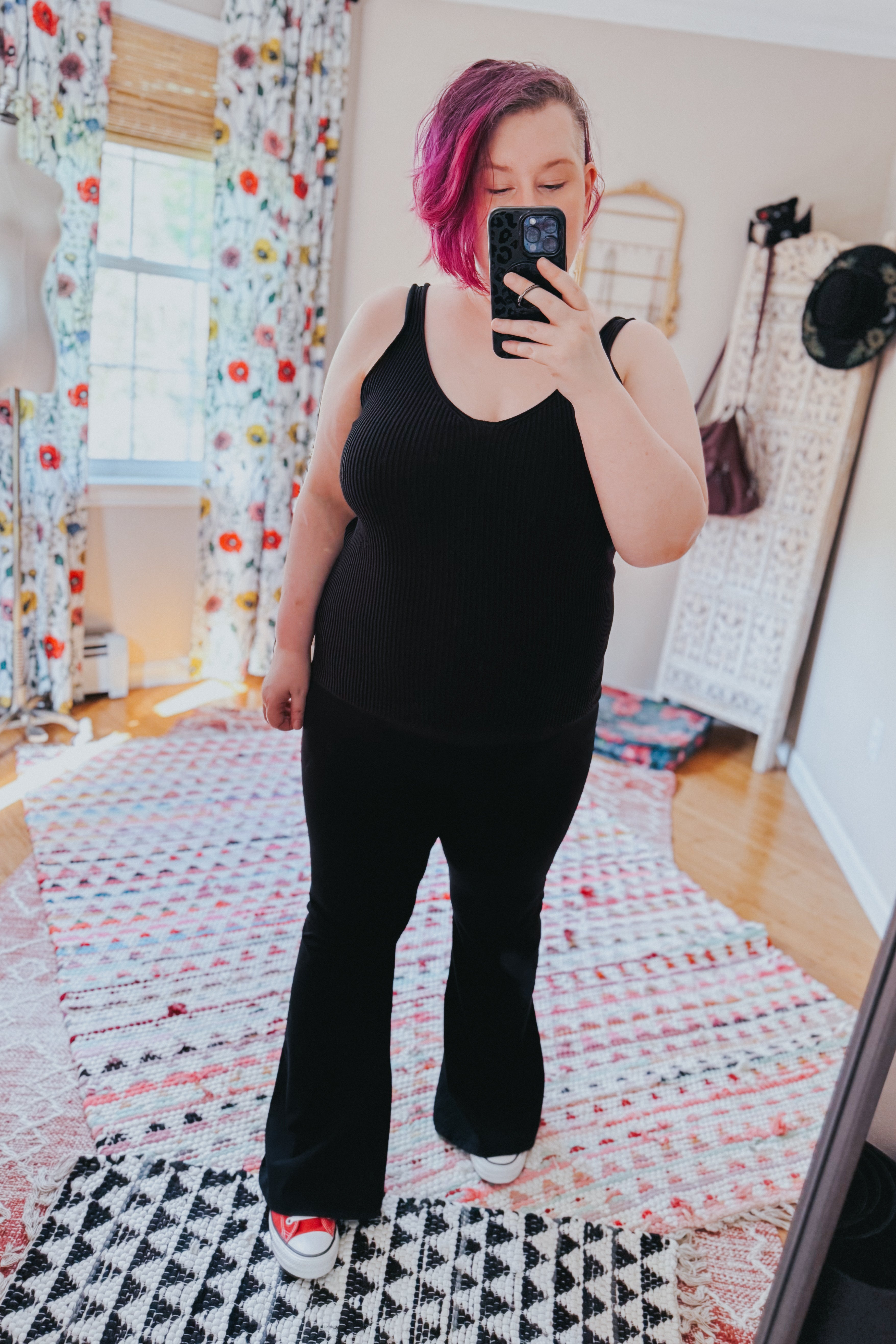 Woman with pink hair wearing a black ribbed tank top and black pants taking a mirror selfie indoors