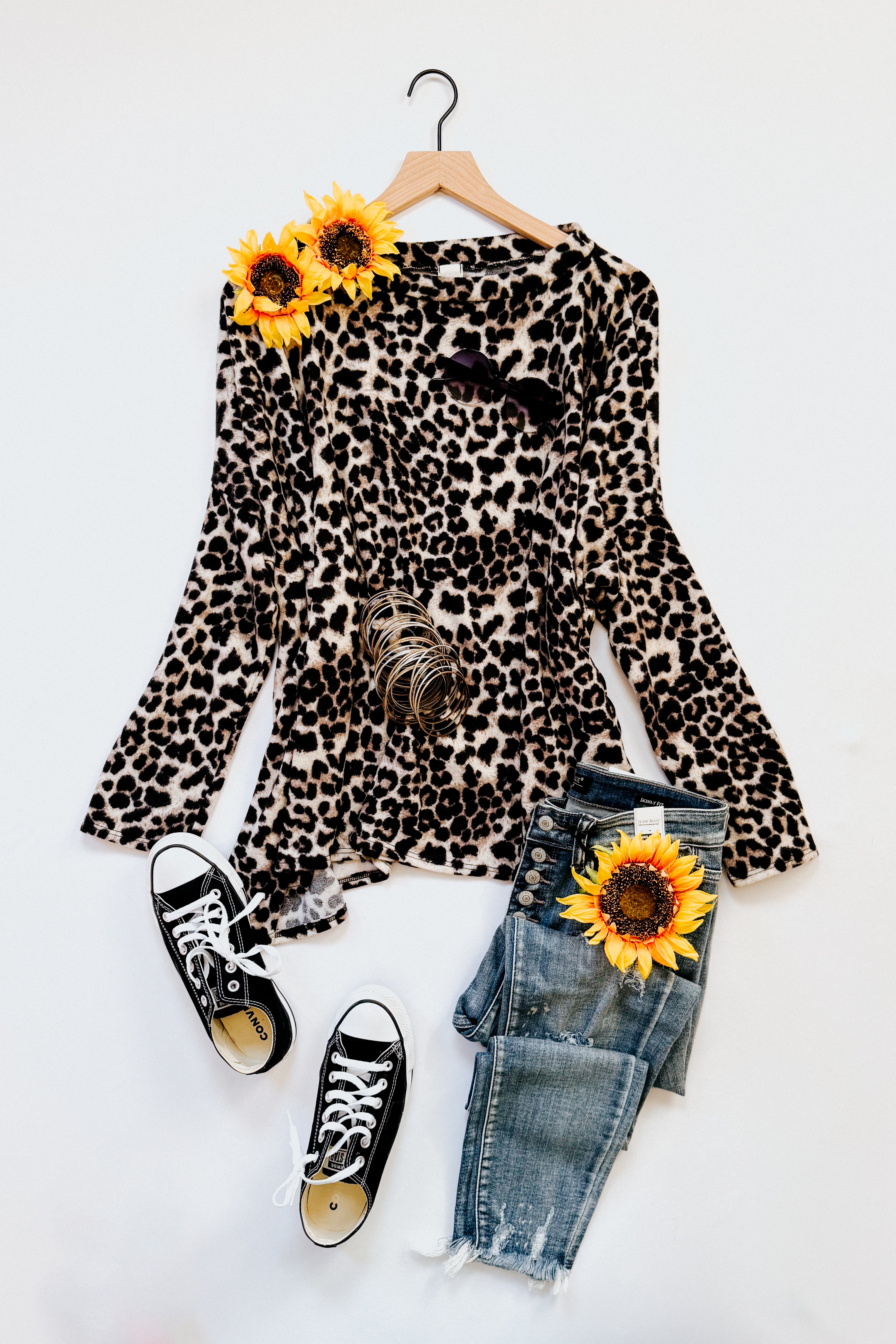 Leopard print long sleeve top with bell sleeves styled with distressed blue jeans, black sneakers, and sunflowers on white background