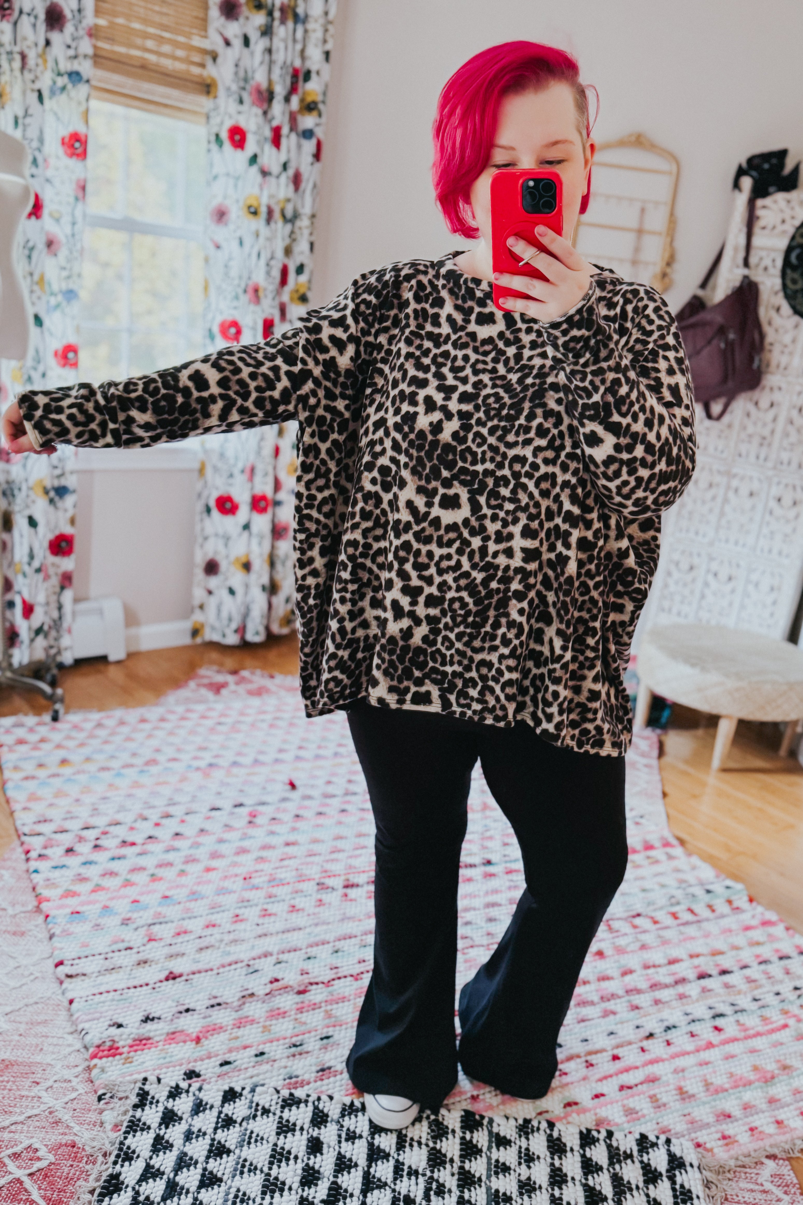 Woman with bright pink hair wearing animal print long sleeve top and black flared pants taking a mirror selfie indoors