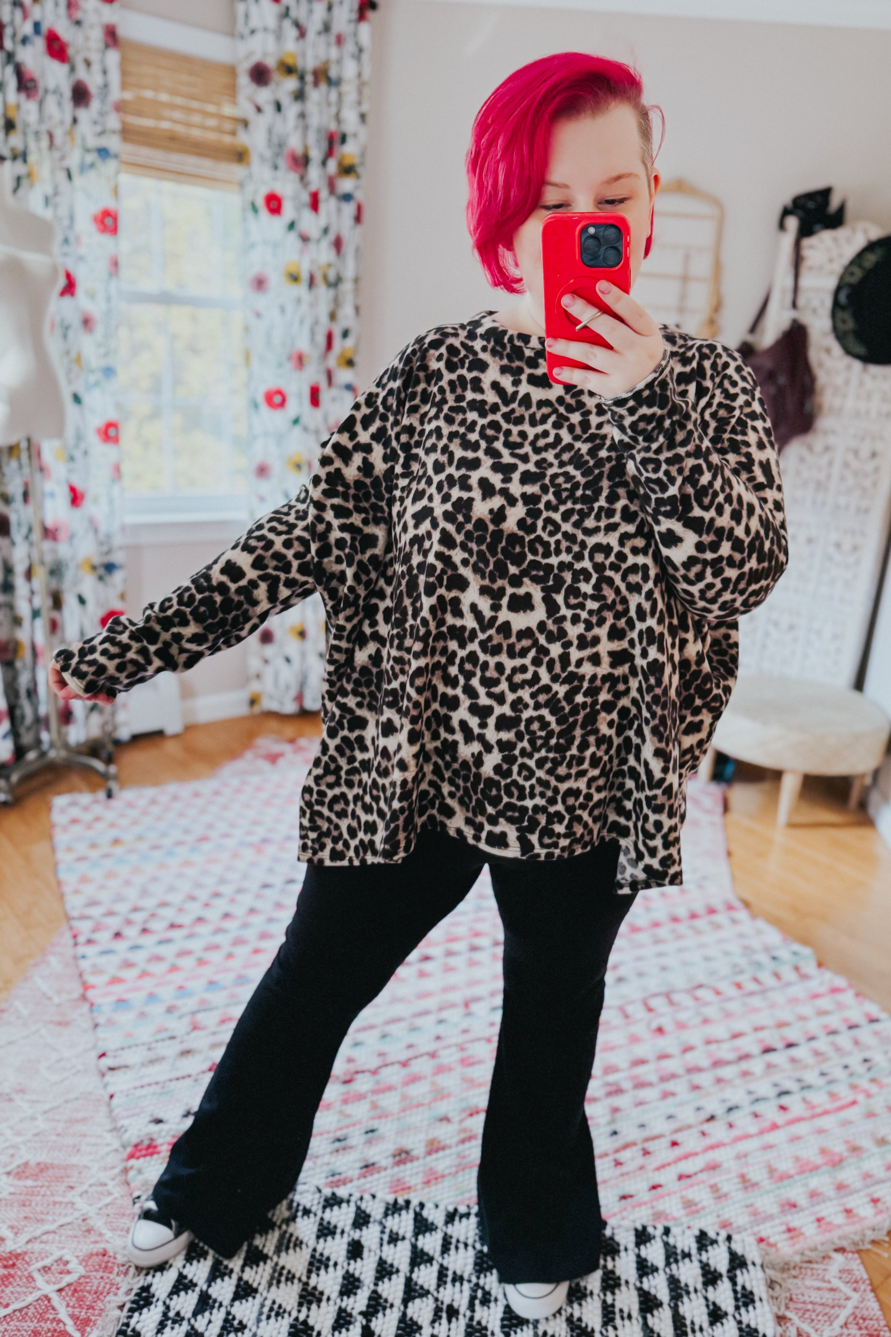 Woman with bright pink hair wearing oversized animal print long sleeve top and black pants taking selfie indoors