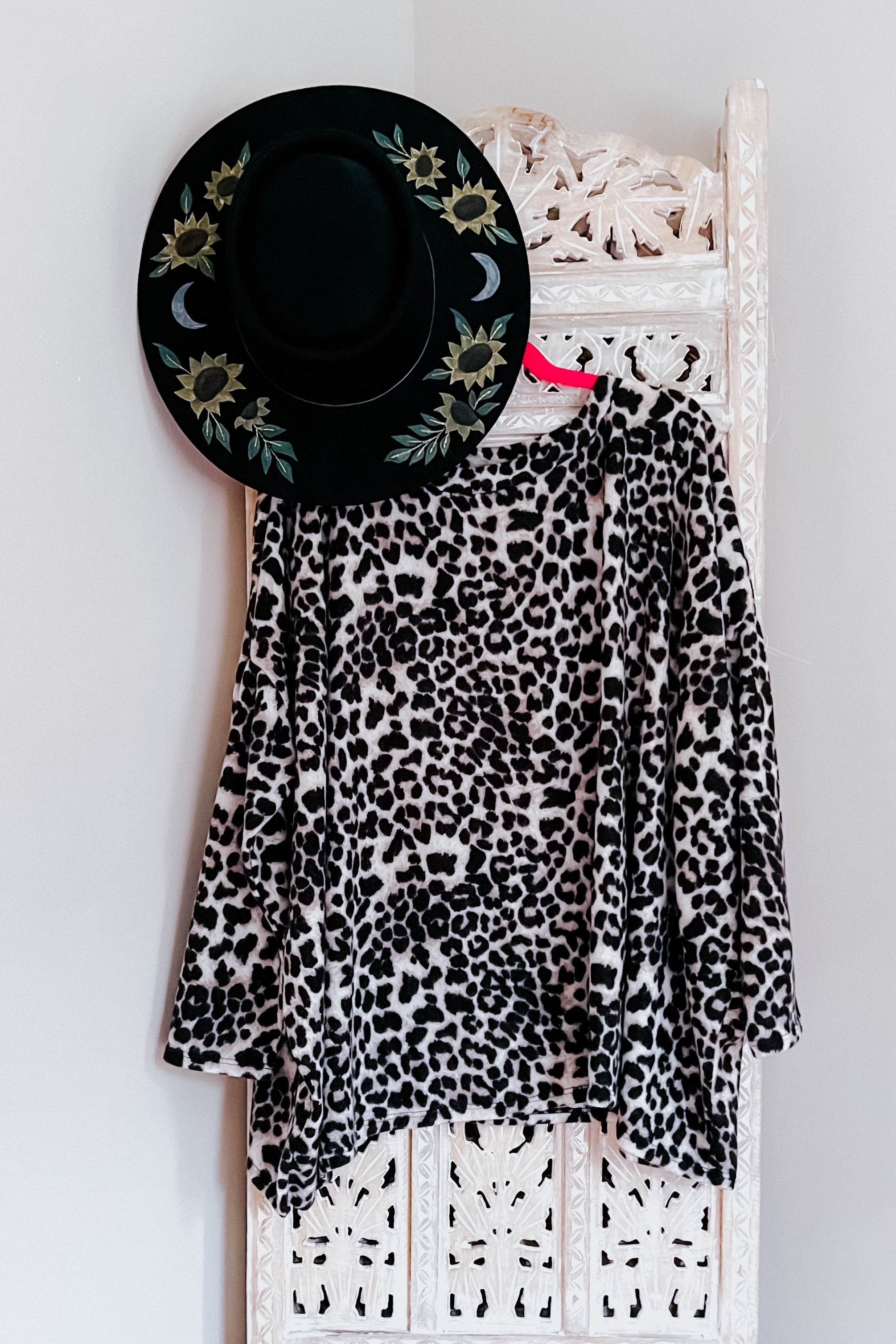 Black and white animal print long sleeve top hanging on carved white wooden panel with black floral hat above