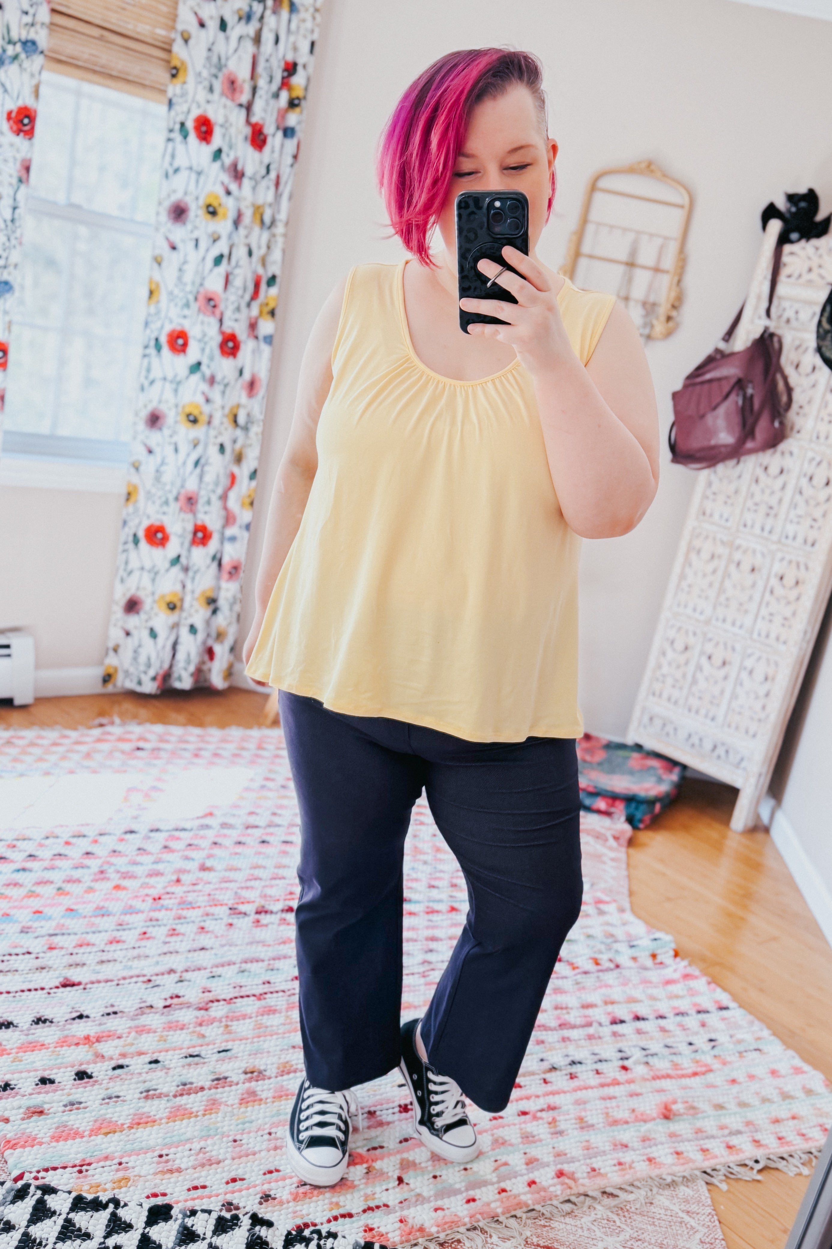Woman with short pink hair wearing sleeveless yellow top and navy cropped pants taking mirror selfie indoors