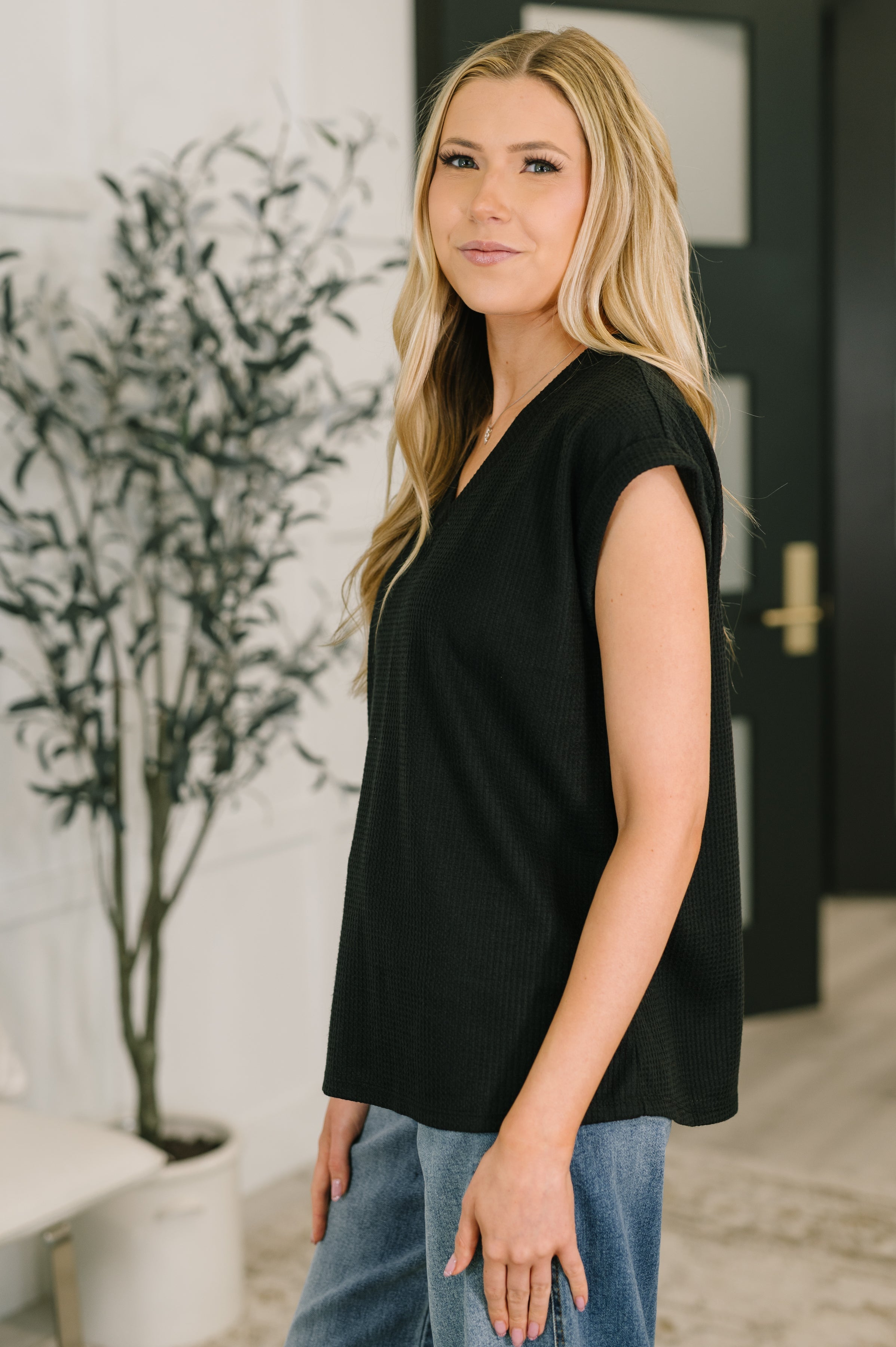 Woman wearing black sleeveless V-neck tee paired with blue jeans standing indoors near plant and door