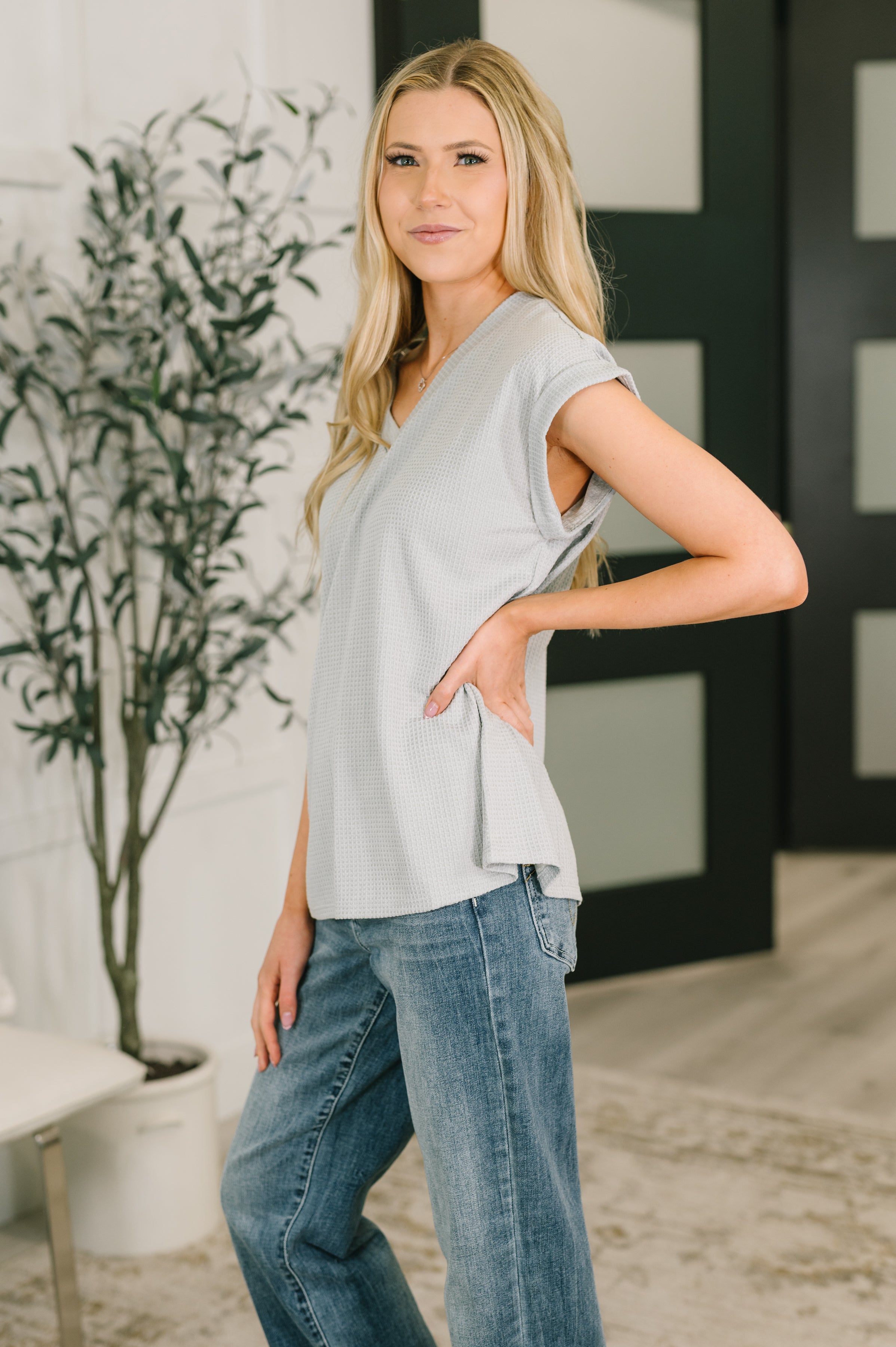 Woman wearing a relaxed pearl grey V-neck tee with rolled sleeves and casual blue jeans posing indoors near plant and door