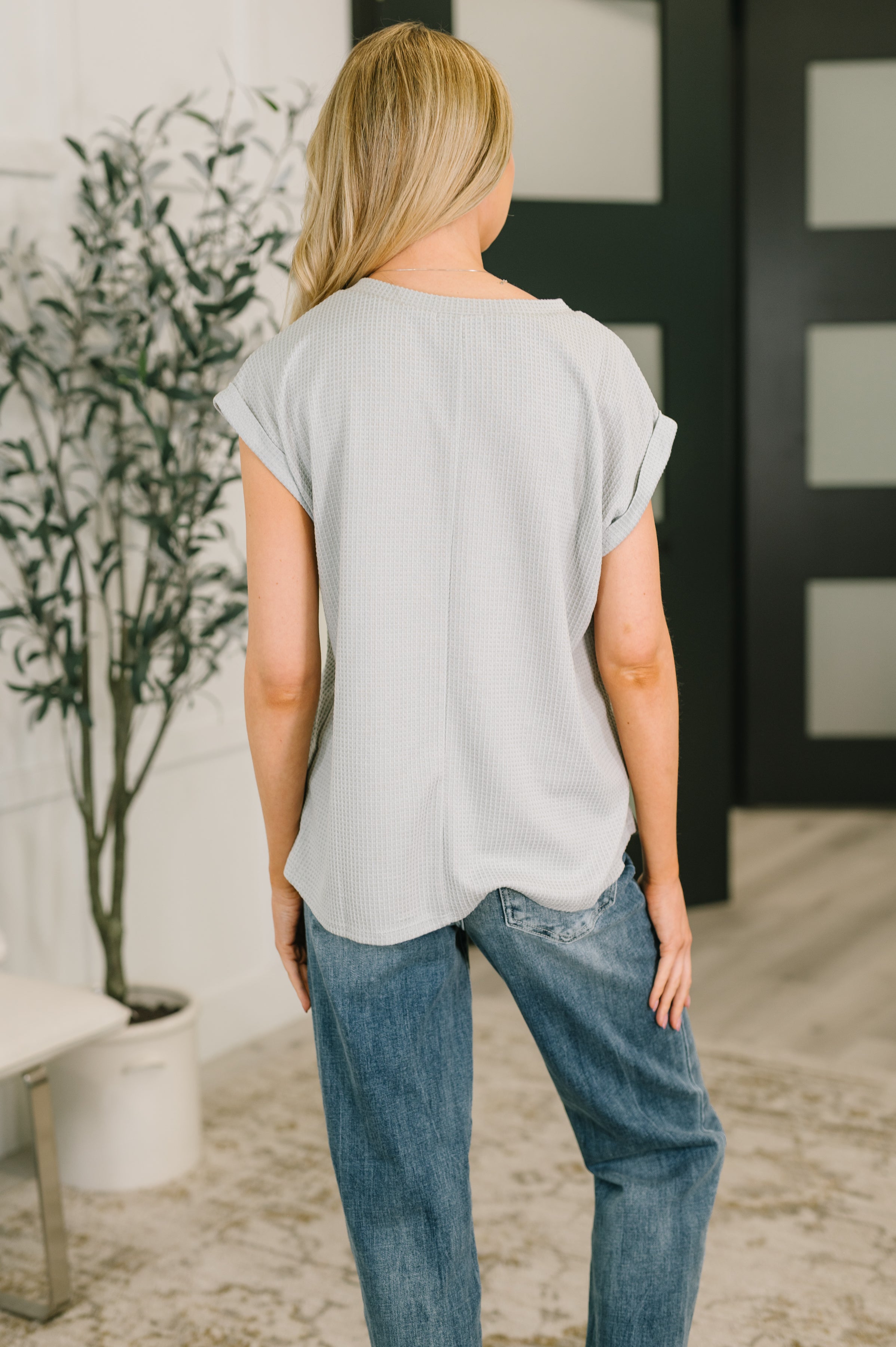 Woman seen from behind wearing pearl grey relaxed fit V-neck top with rolled sleeves and blue jeans indoors
