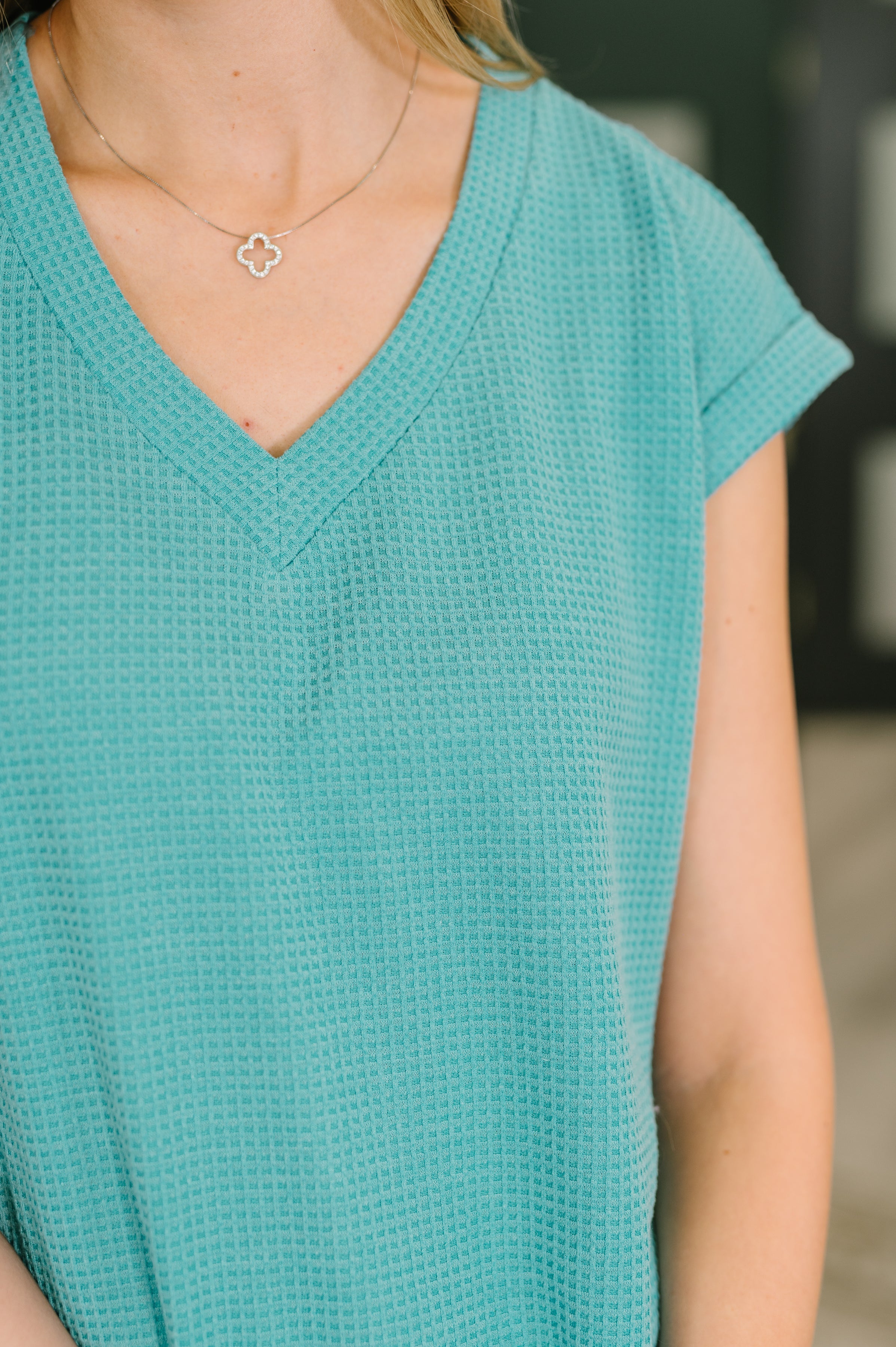 Close-up of woman wearing teal textured V-neck tee with short cuffed sleeves and delicate silver clover necklace indoors