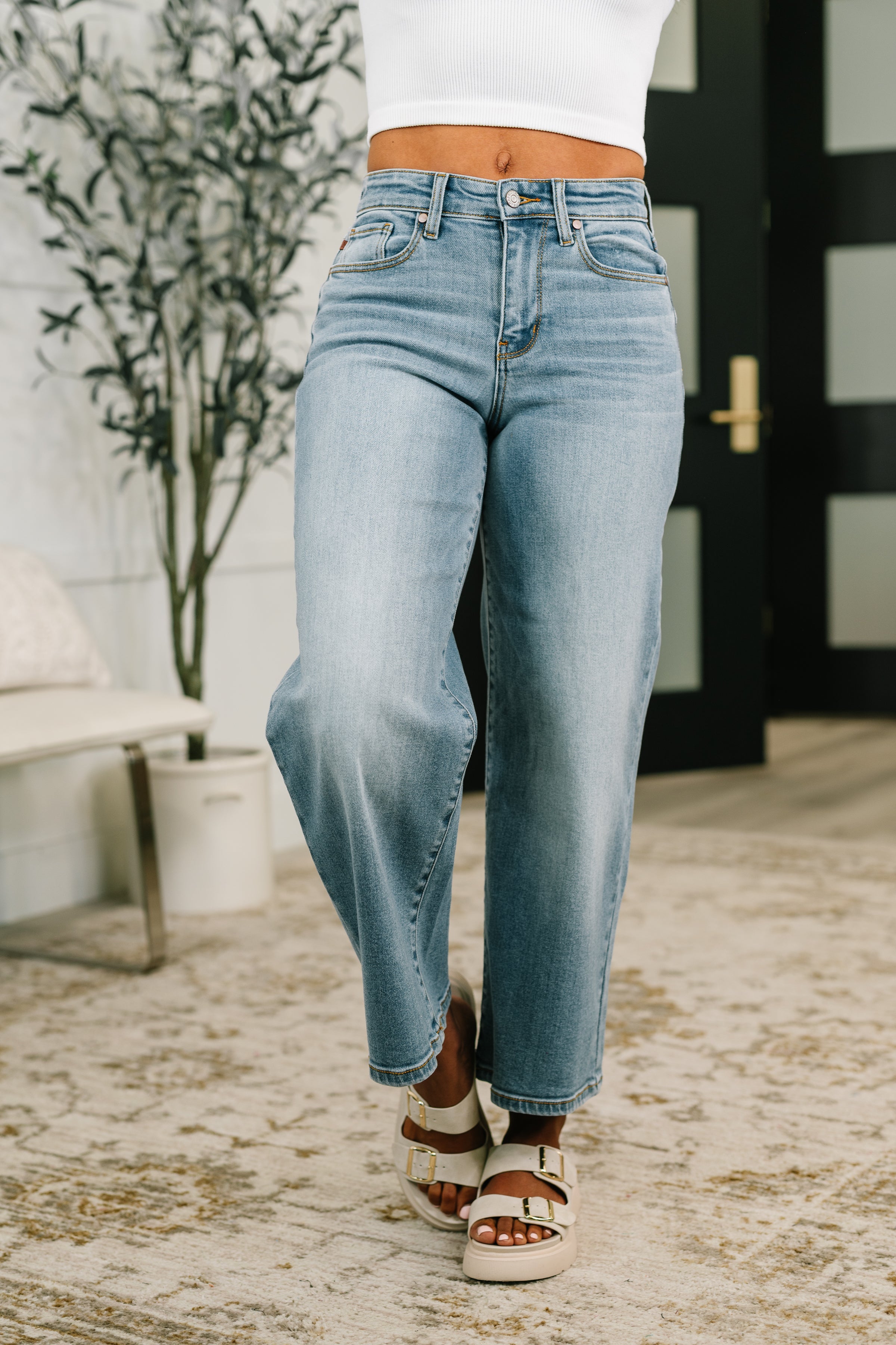 Woman wearing mid rise wide leg blue jeans and white buckle sandals standing indoors on patterned rug