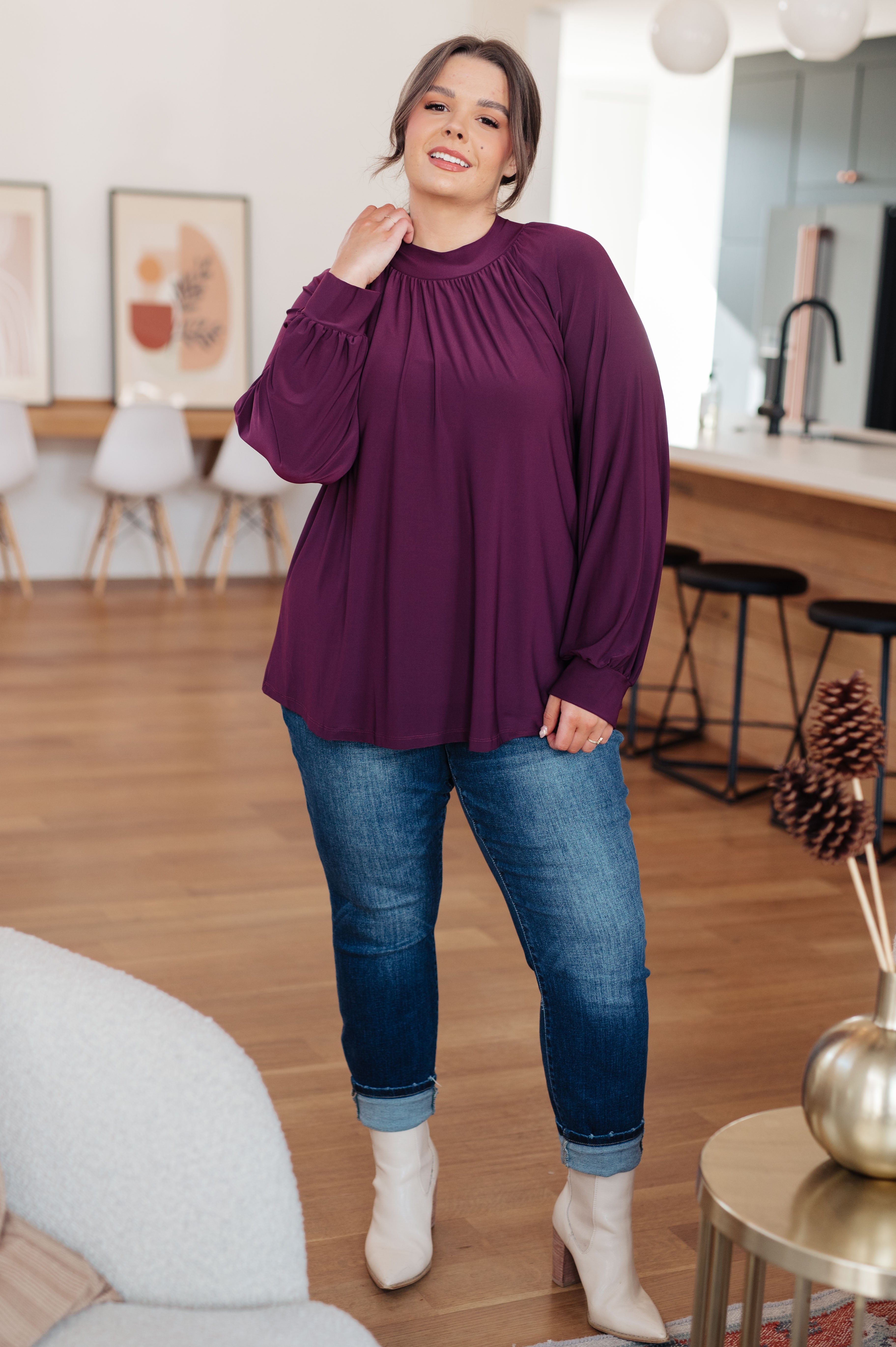 Plus size woman wearing a flowing plum mock neck blouse with long balloon sleeves paired with cuffed jeans and white boots