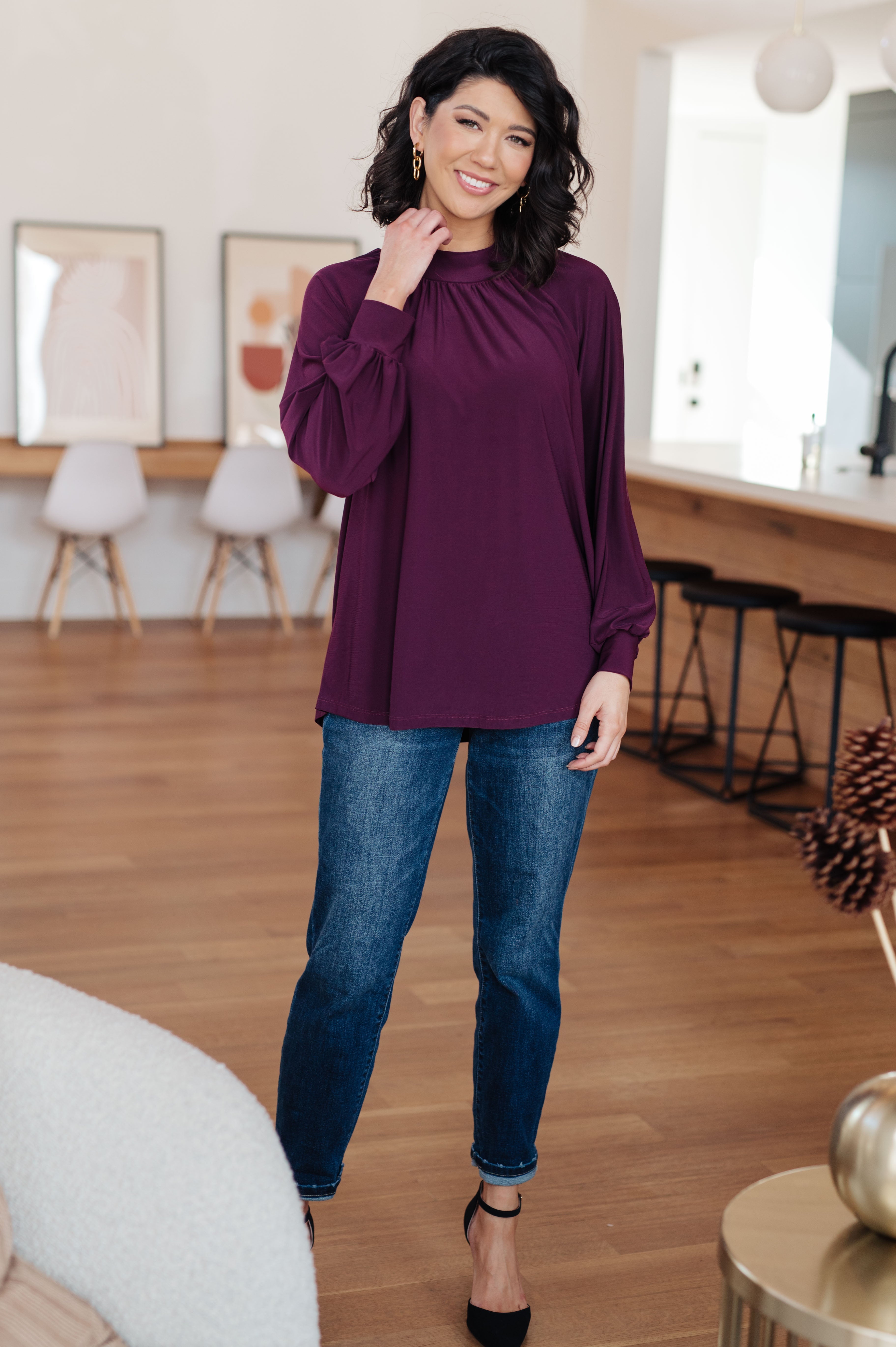 Woman wearing a plum mock neck blouse with long puffed sleeves paired with dark blue jeans in a cozy living space