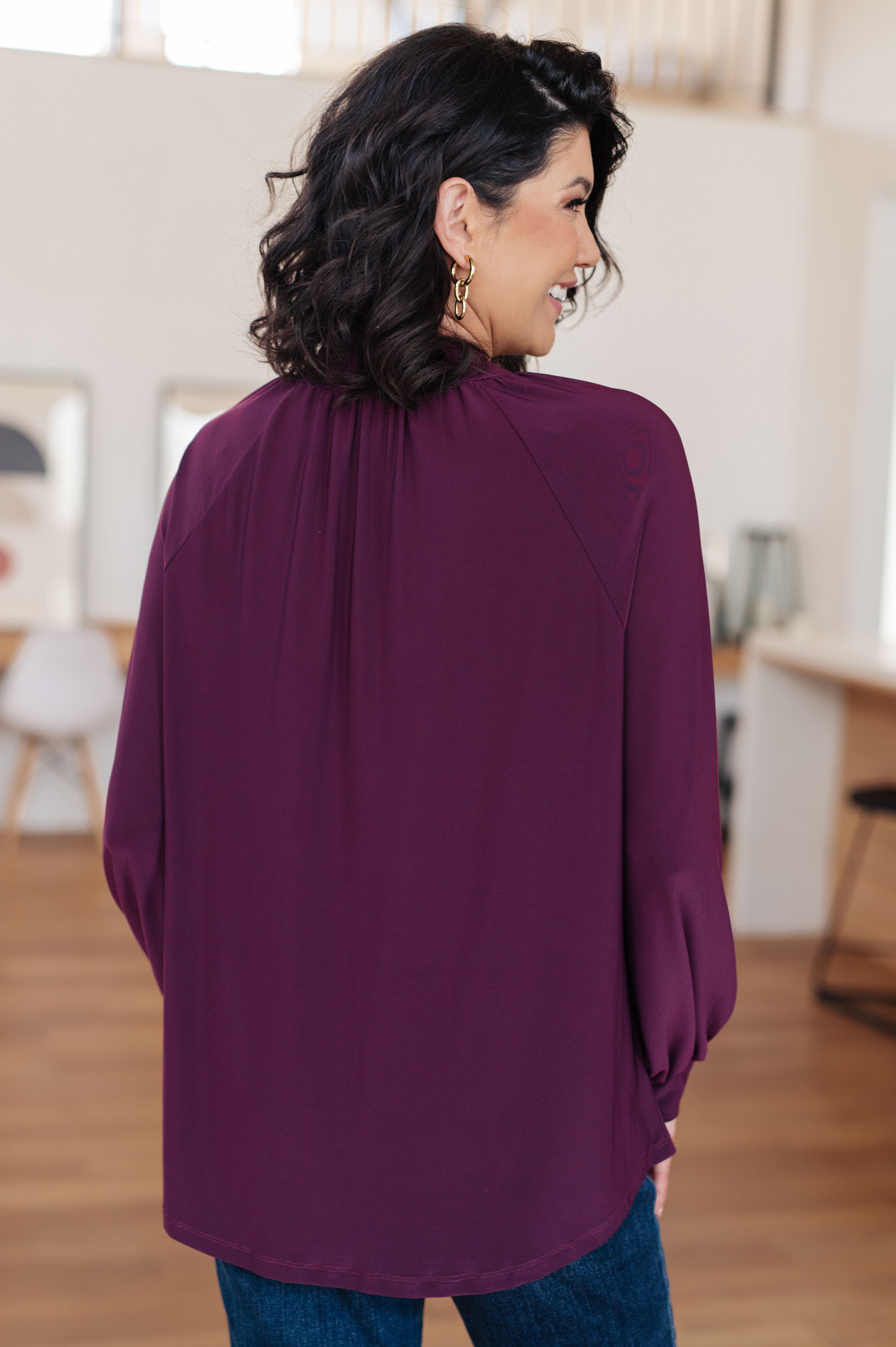 Woman wearing a deep purple mock neck blouse with long sleeves and subtle pleats at the back indoors