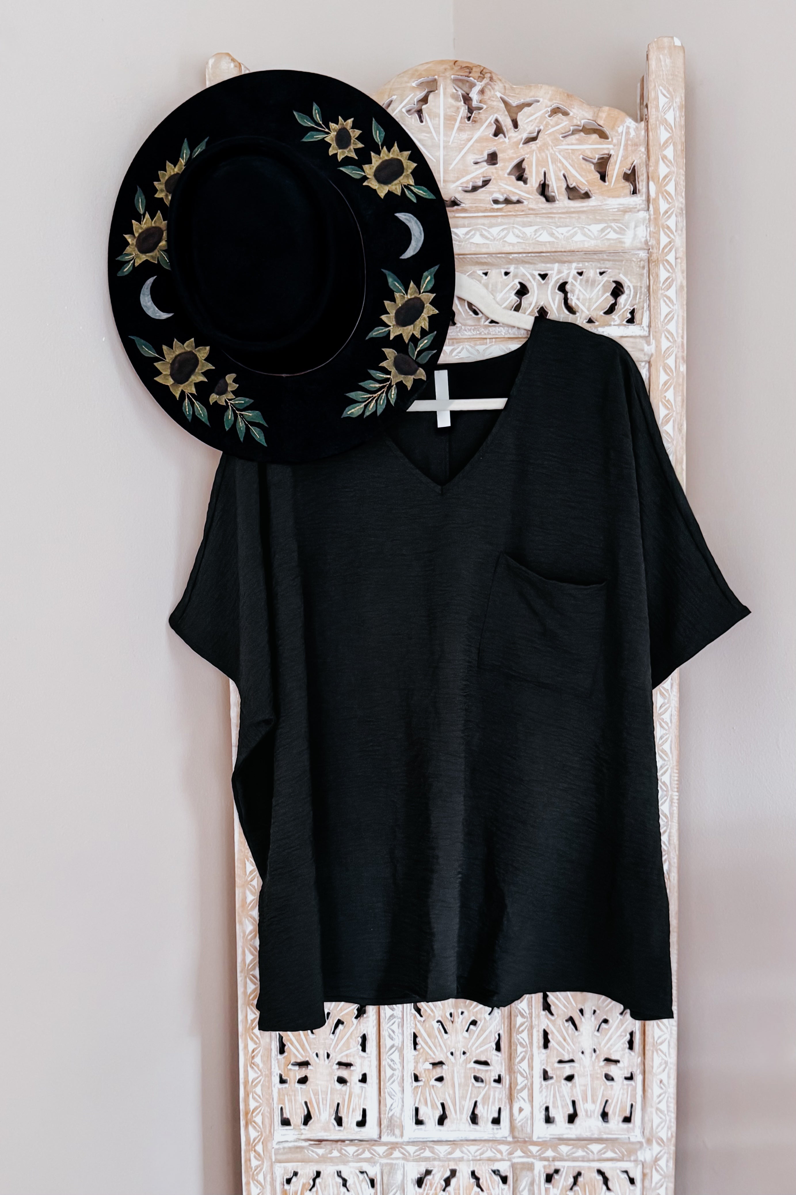 Black relaxed-fit V-neck top with short sleeves and chest pocket hanging on carved wooden chair with embroidered black hat