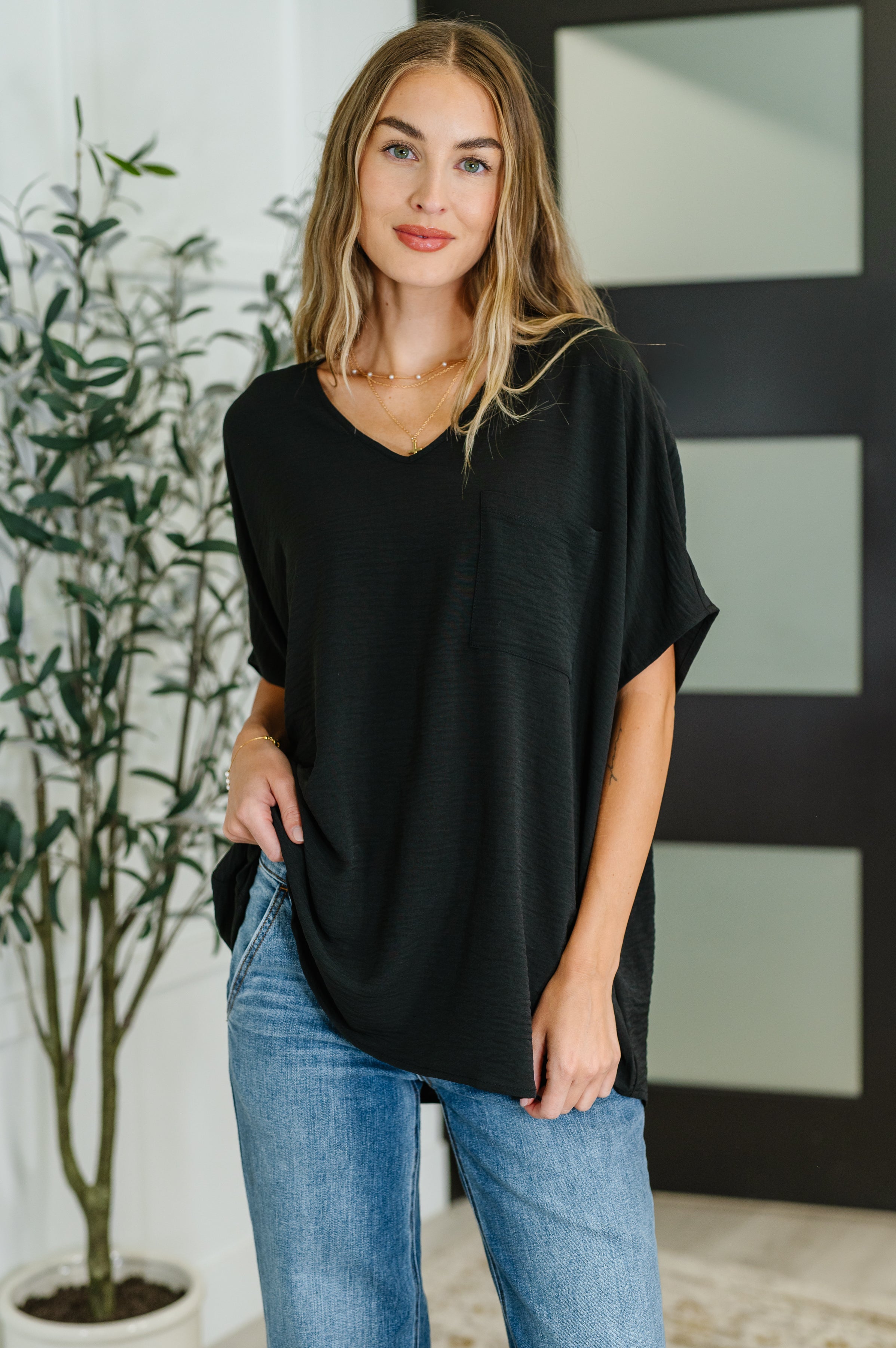 Young woman wearing relaxed black V-neck top with short sleeves and front pocket paired with casual blue jeans indoors