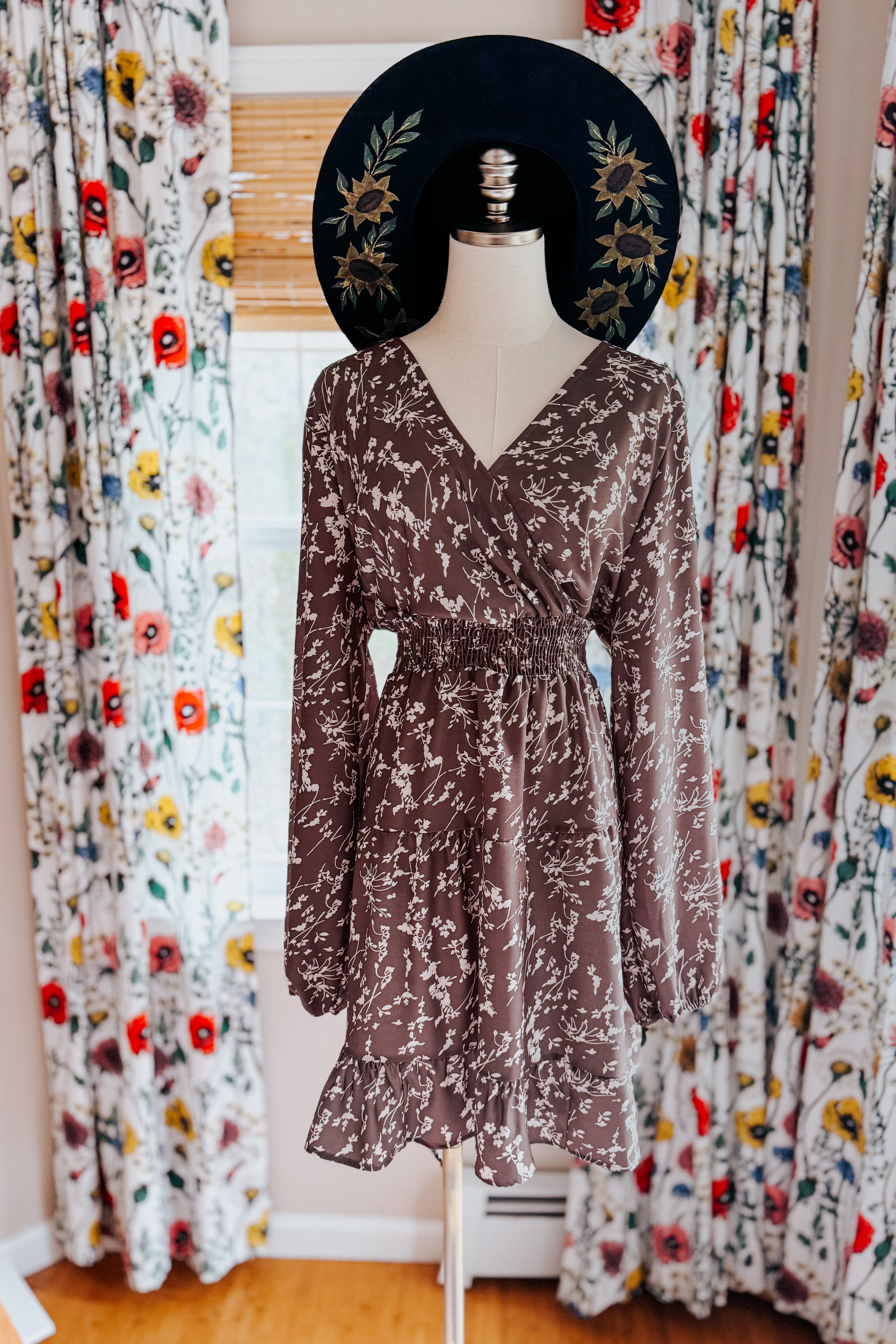 Mannequin wearing cocoa brown floral dress with smocked waist, long sleeves, V-neck, and ruffled hem standing indoors