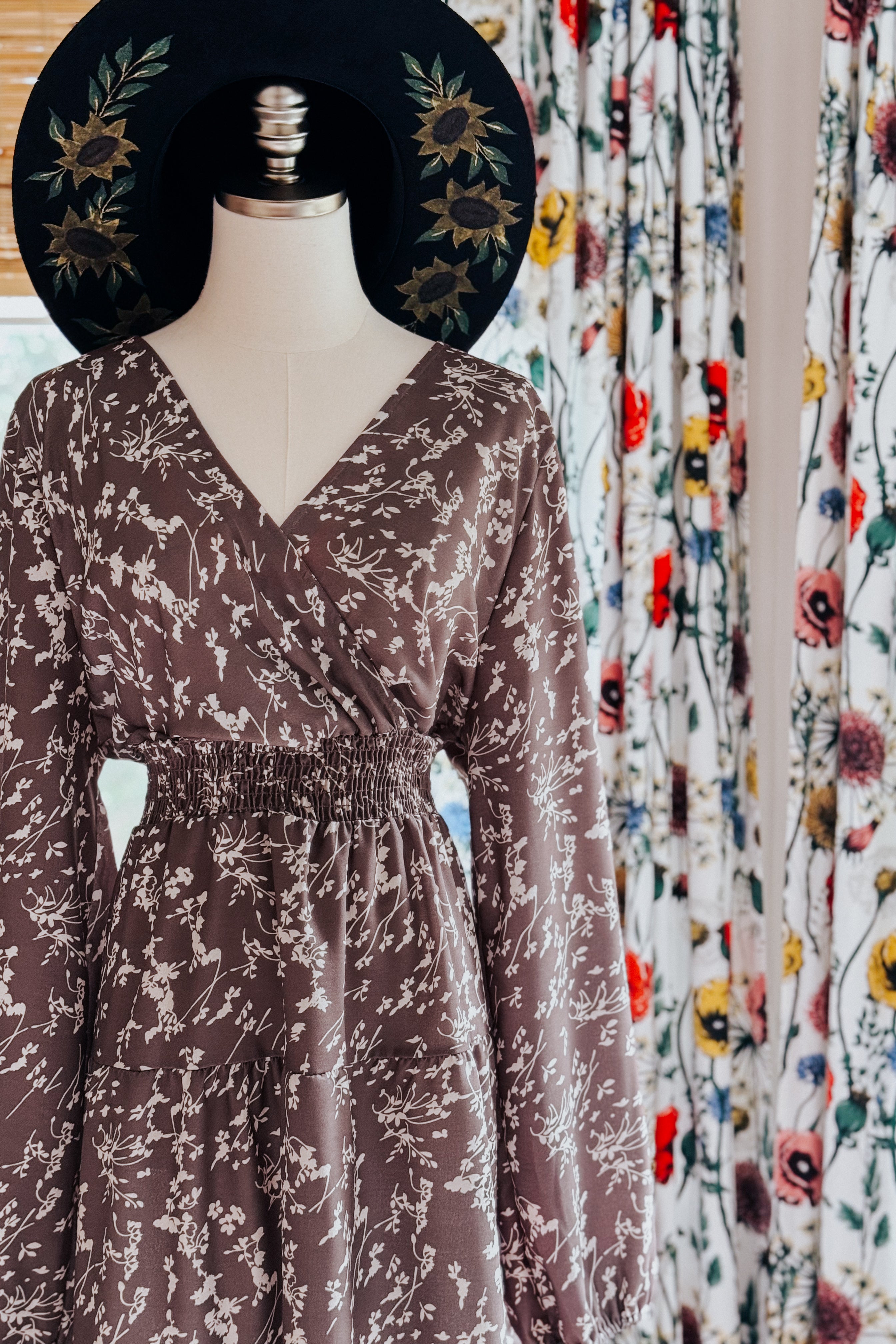 Mannequin dressed in cocoa brown floral wrap dress with smocked waist and long sleeves, accessorized with black sunflower-embroidered hat