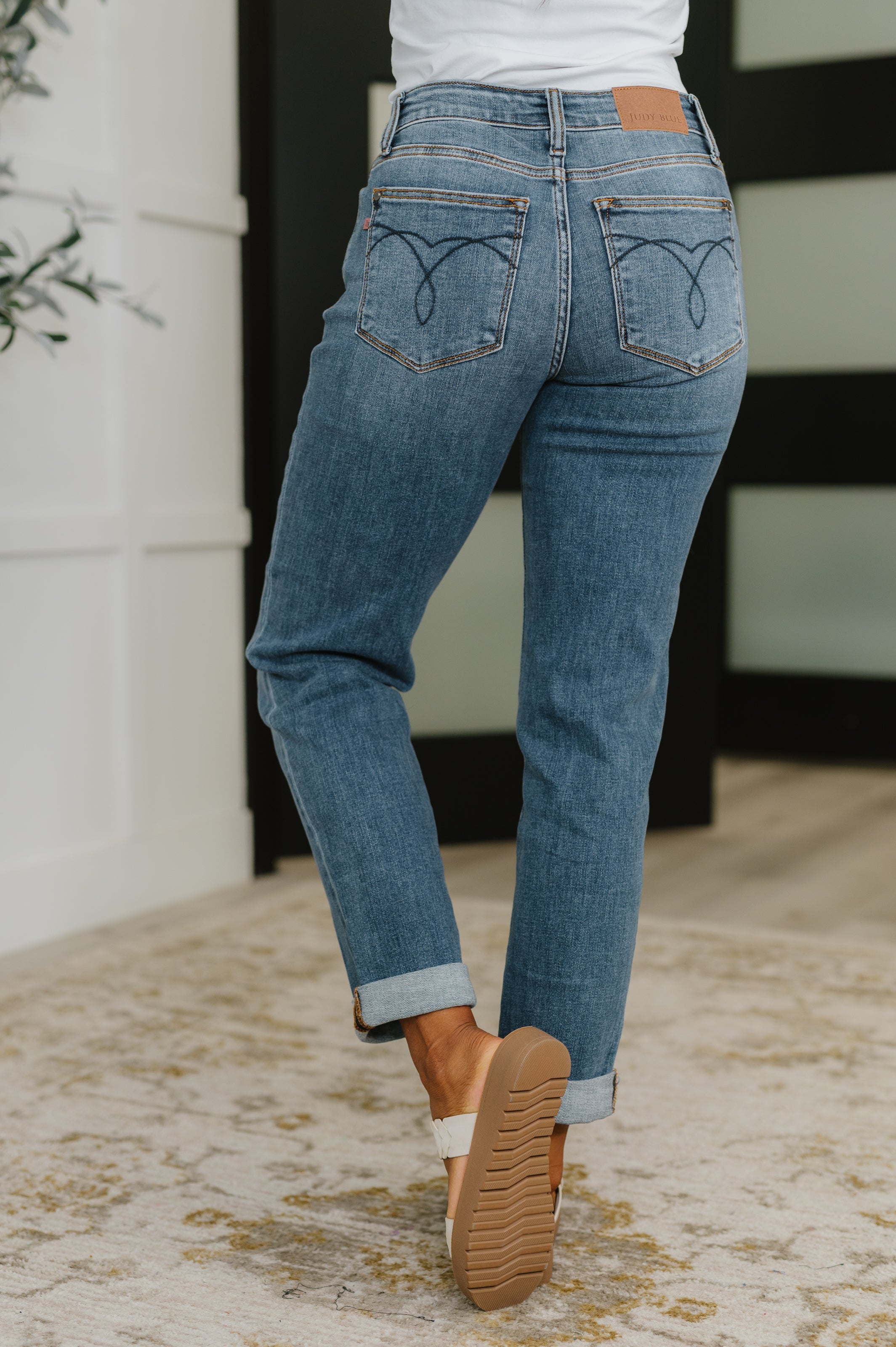 Back view of woman wearing blue cuffed boyfriend jeans with a white top and tan shoes indoors