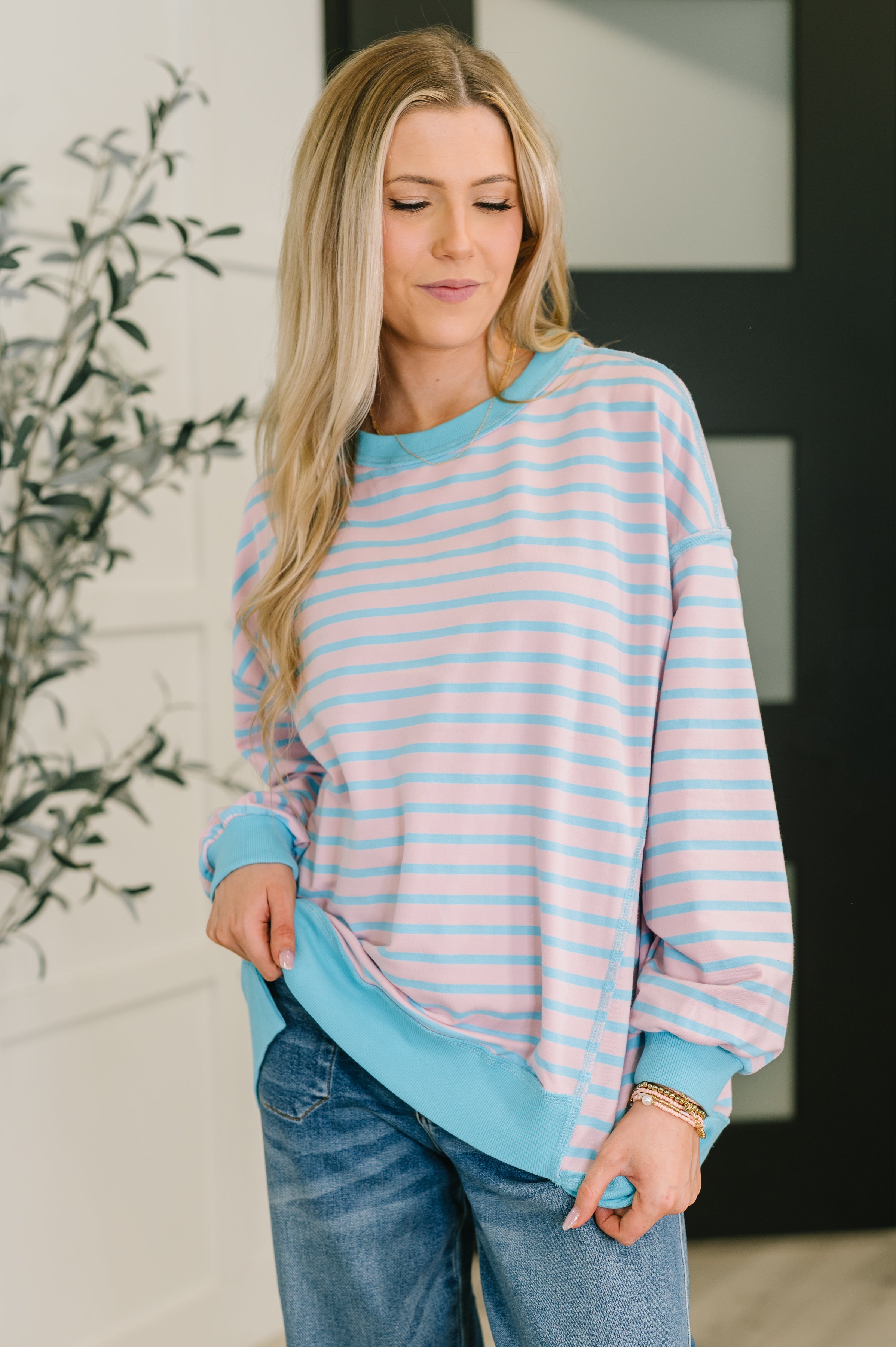 Woman wearing pastel blue and pink striped drop shoulder long sleeve pullover paired with casual blue jeans indoors