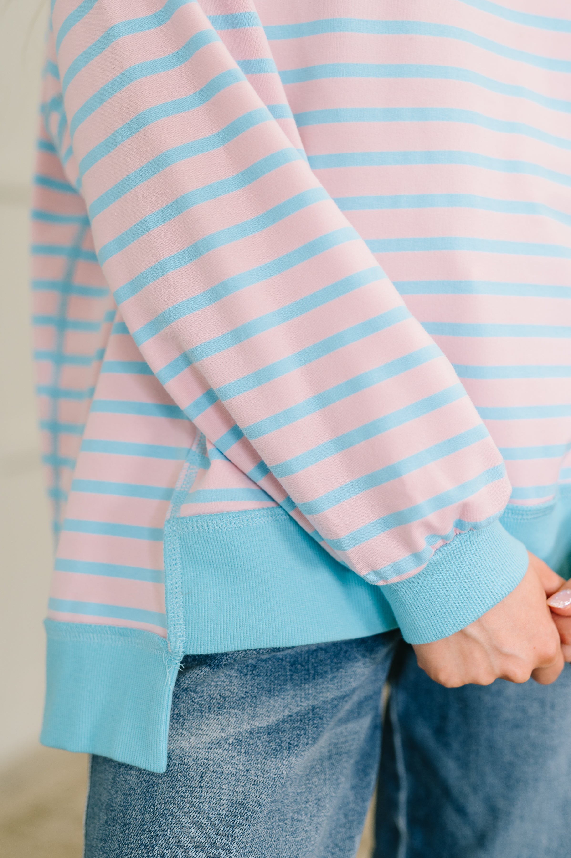 Close-up of person wearing pastel pink and blue striped long sleeve pullover with ribbed cuffs and hem over jeans