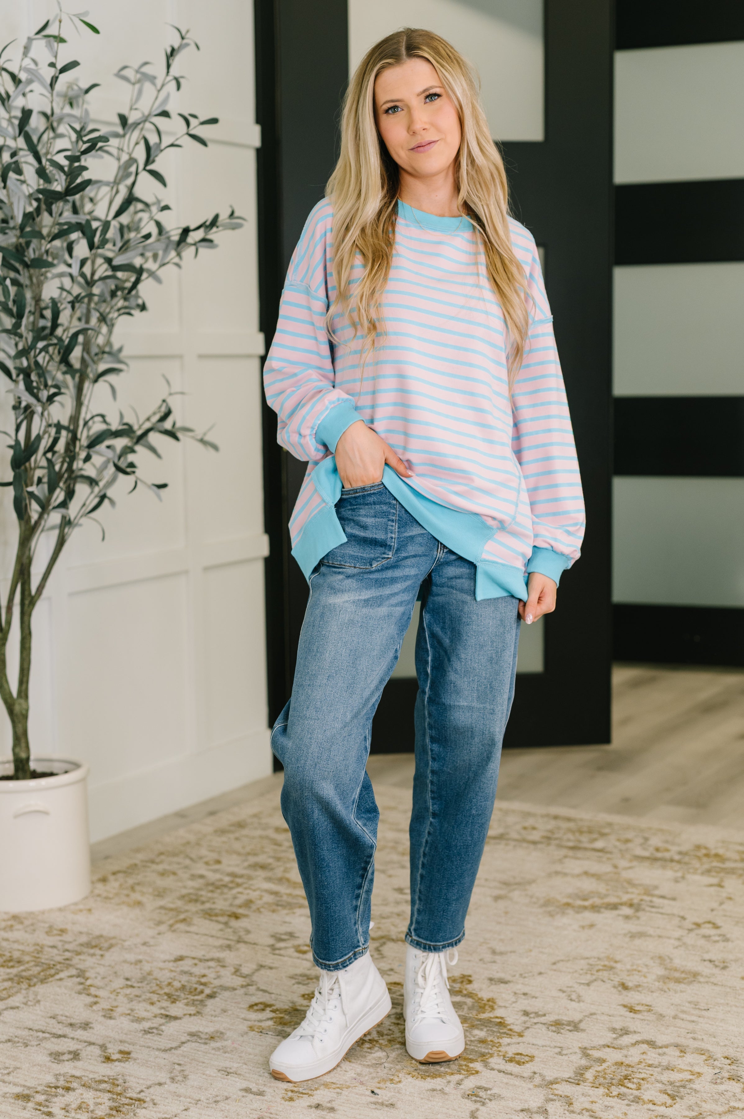 Woman standing indoors wearing pastel blue and pink striped drop shoulder long sleeve pullover with jeans and white boots