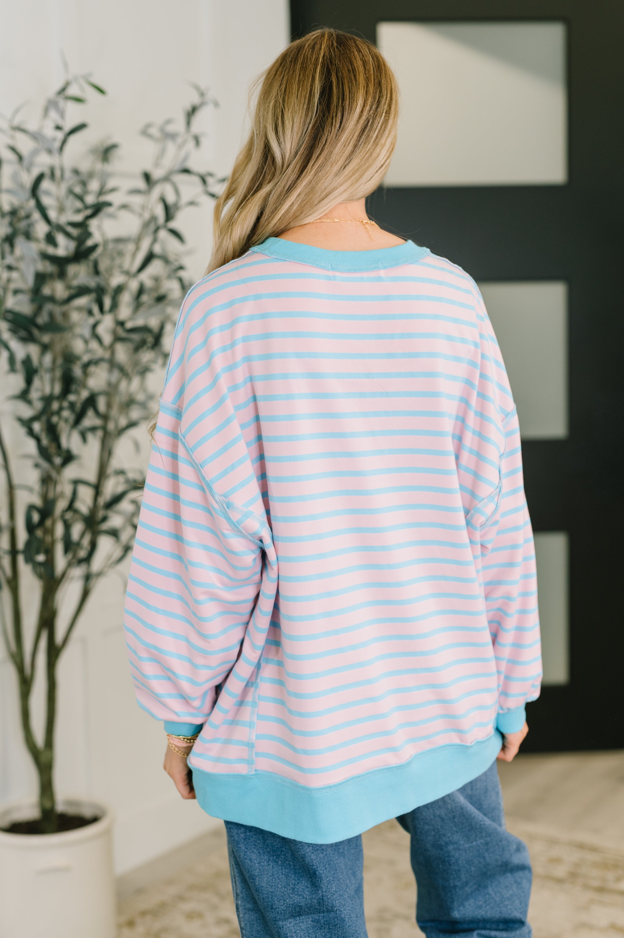 Woman wearing pastel blue and pink striped drop shoulder long sleeve pullover viewed from the back indoors