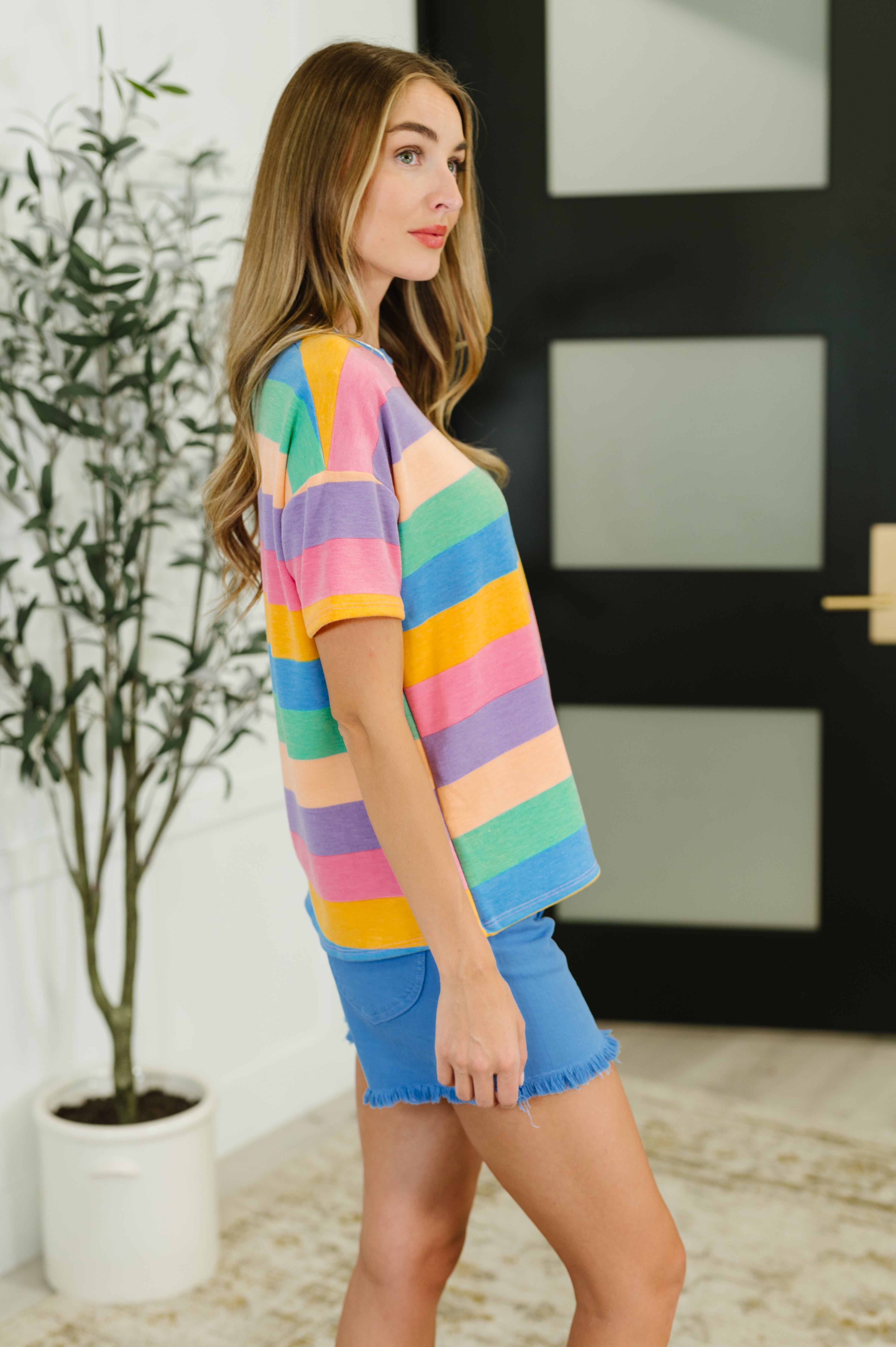 Side view of woman wearing colorful striped drop shoulder top with blue frayed hem shorts indoors