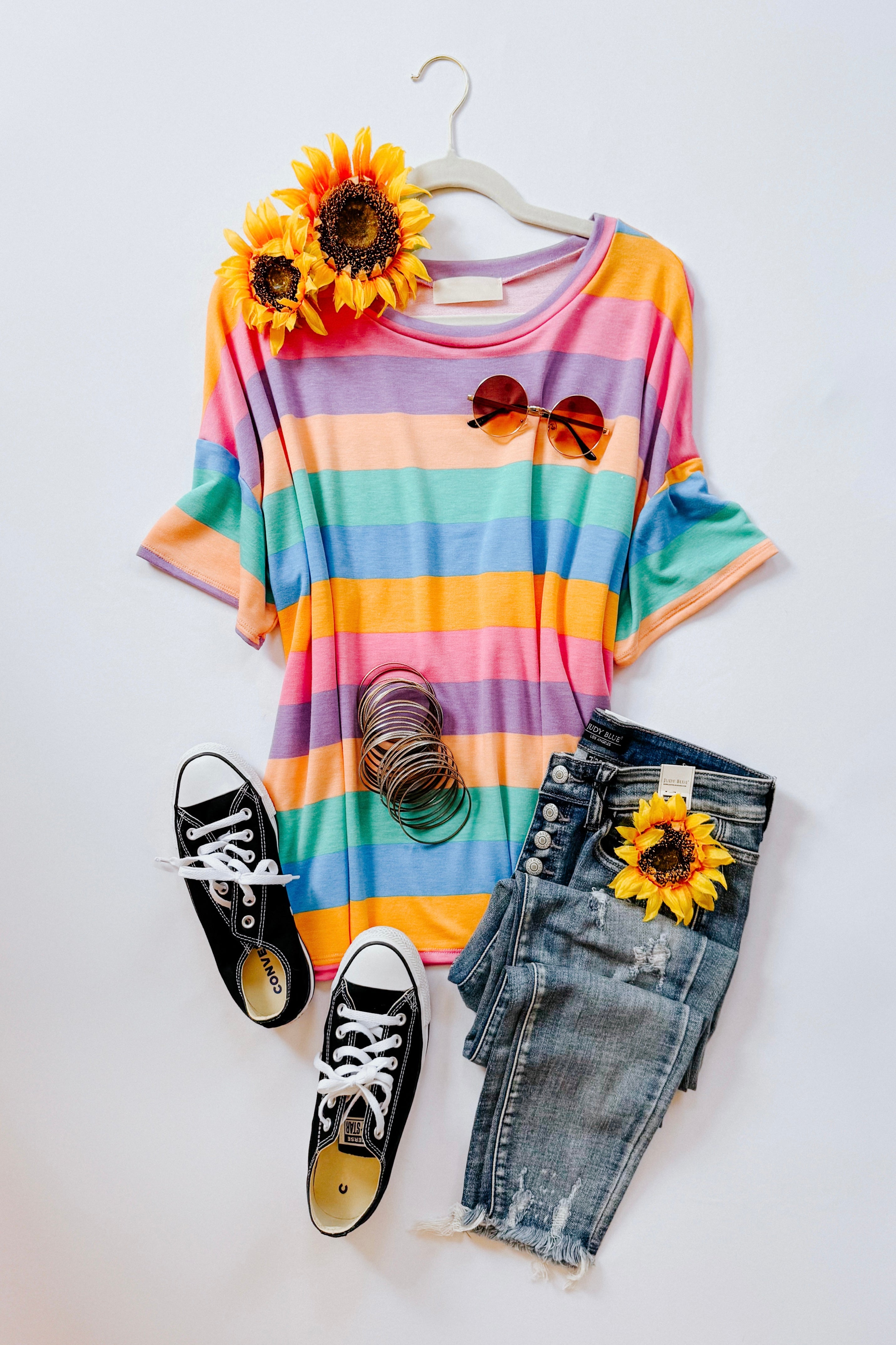 Vibrant striped drop shoulder top paired with distressed denim shorts, black sneakers, sunglasses, and sunflower accents on white background