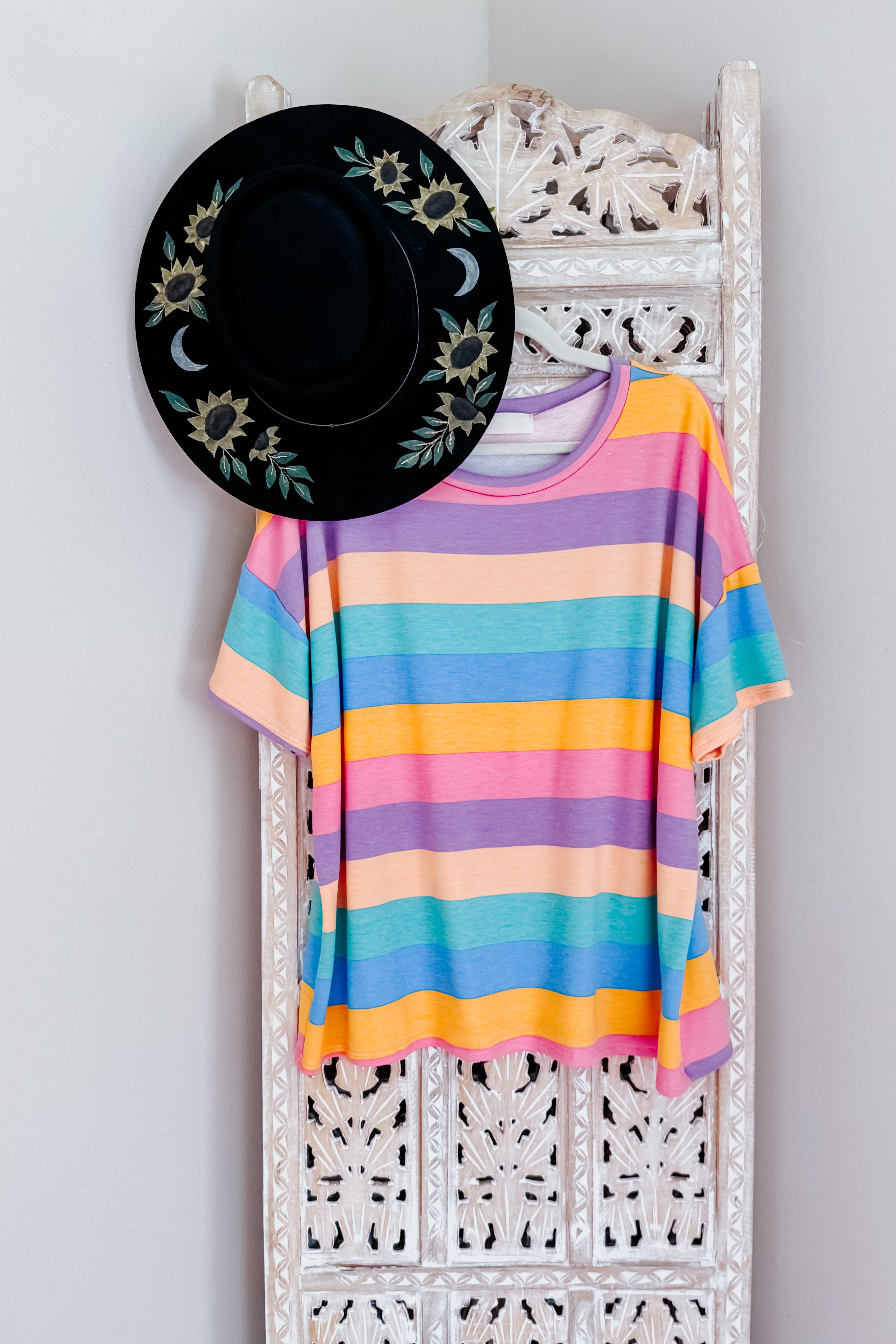 Multicolored striped drop shoulder top hanging on ornate white carved wooden panel with black floral hat