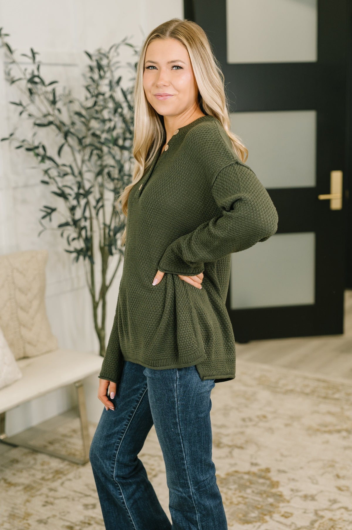 Woman modeling a dark olive sweater knit henley top with a V neckline and relaxed fit styled with blue jeans indoors