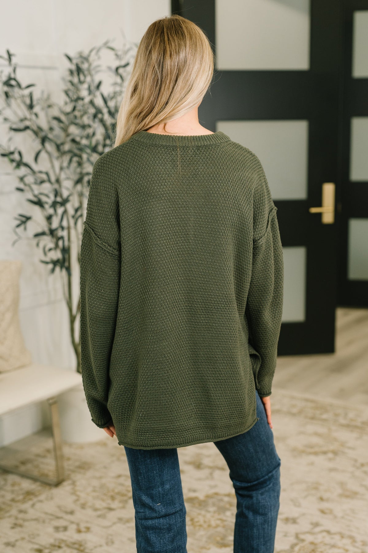 Woman seen from behind wearing a dark olive textured knit top with long sleeves and blue jeans in a softly styled interior