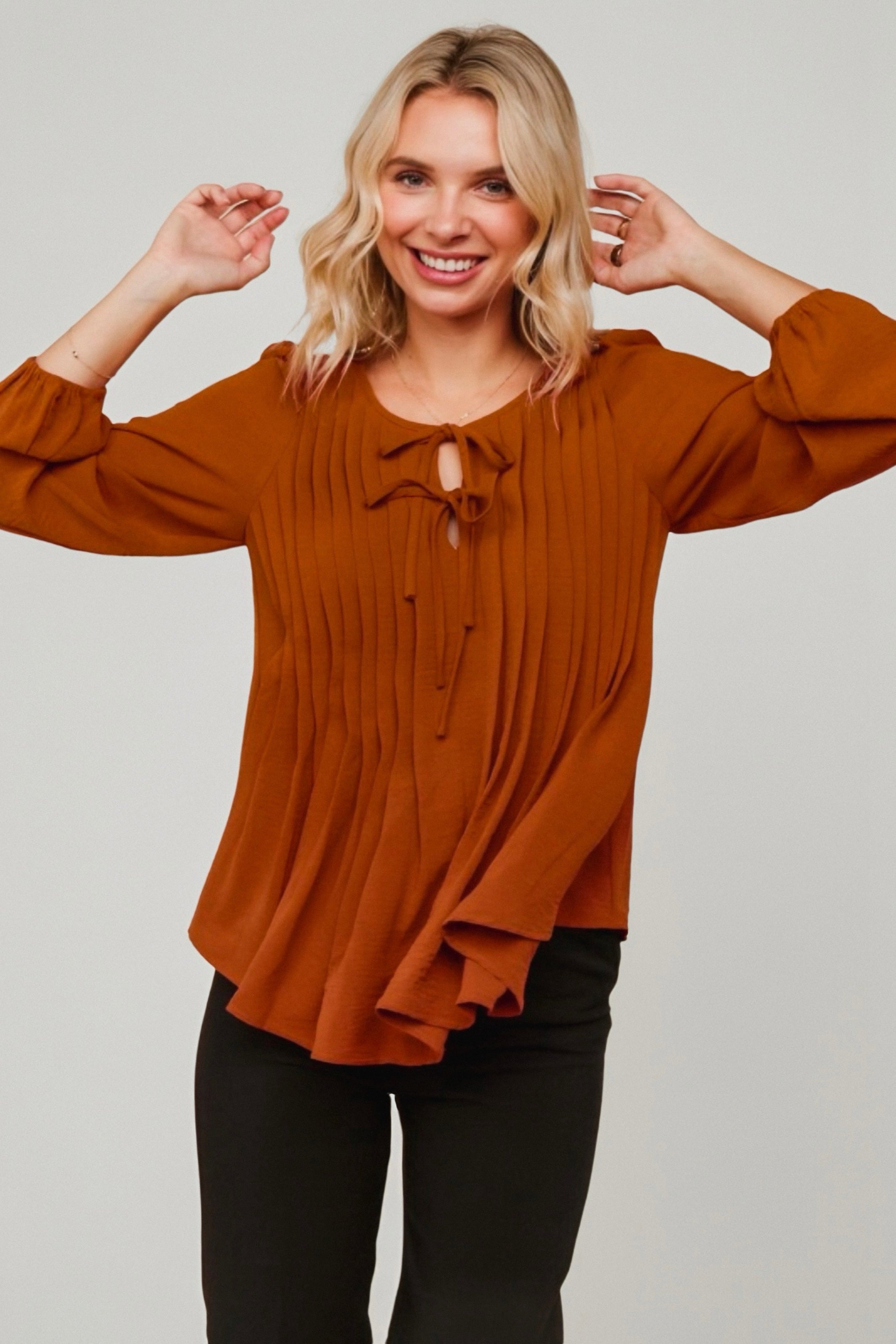 Smiling woman wearing copper pleated blouse with keyhole and tie neckline and balloon sleeves against neutral background