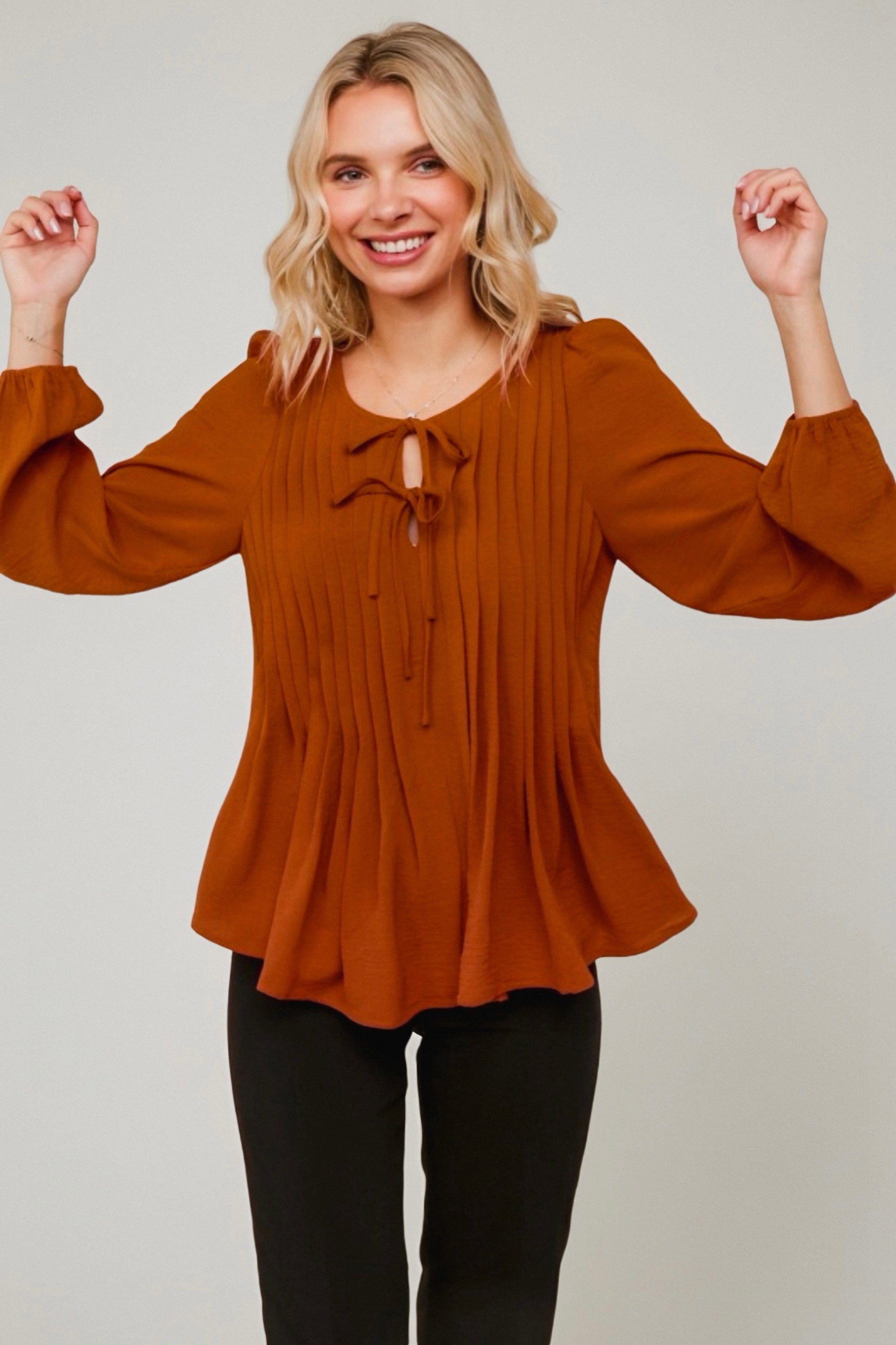Smiling woman wearing copper pleated blouse with long bishop sleeves and tie details paired with black pants against light background