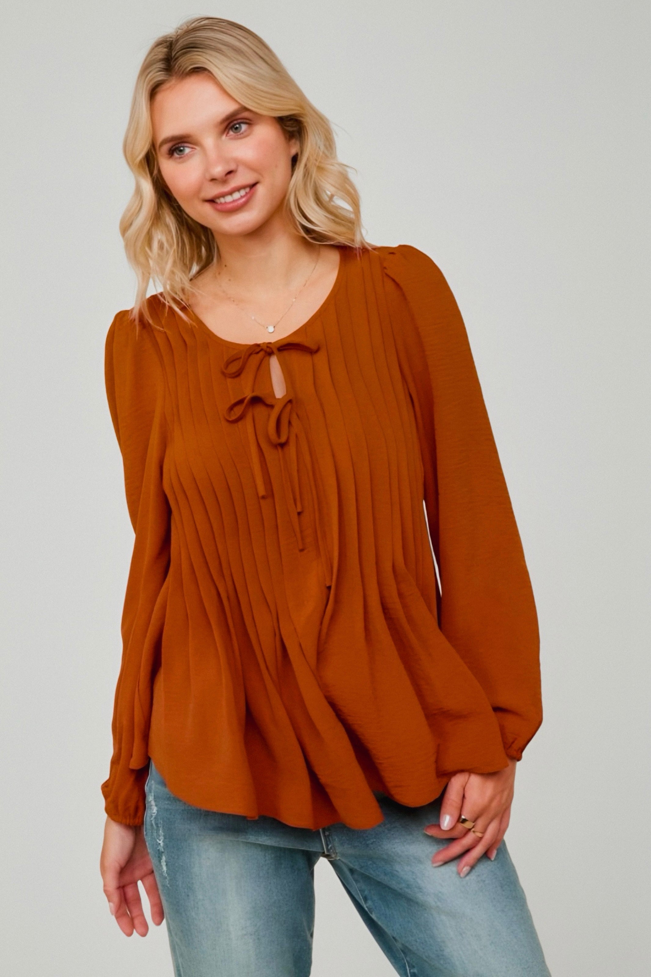 Woman wearing rust-colored pleated blouse with long sleeves and double bow ties at neckline paired with light-wash jeans