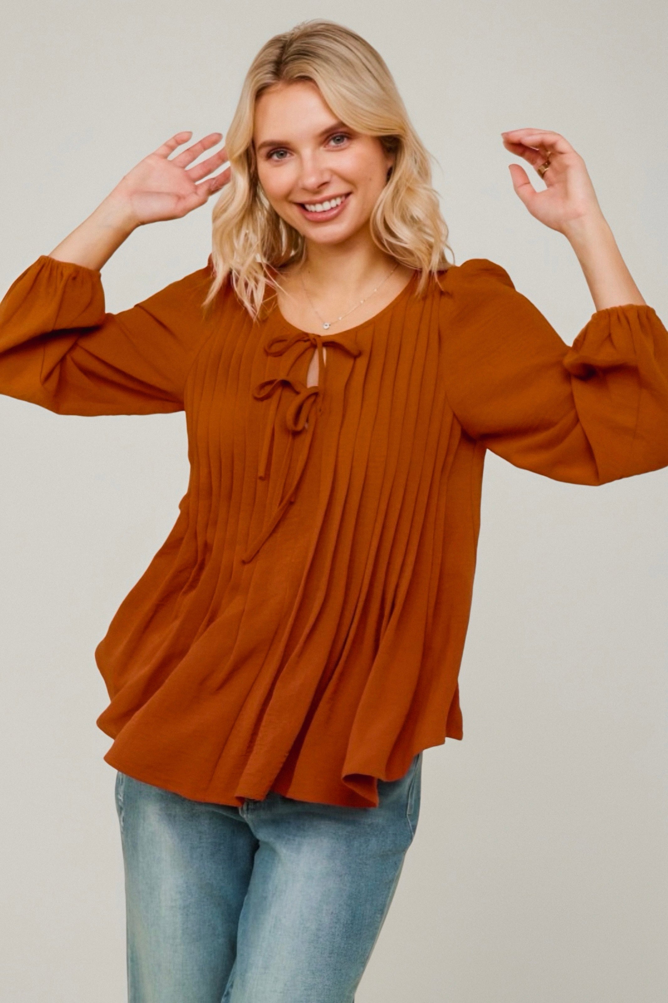 Smiling woman wearing copper pleated blouse with balloon sleeves and front tie detail paired with light-wash jeans