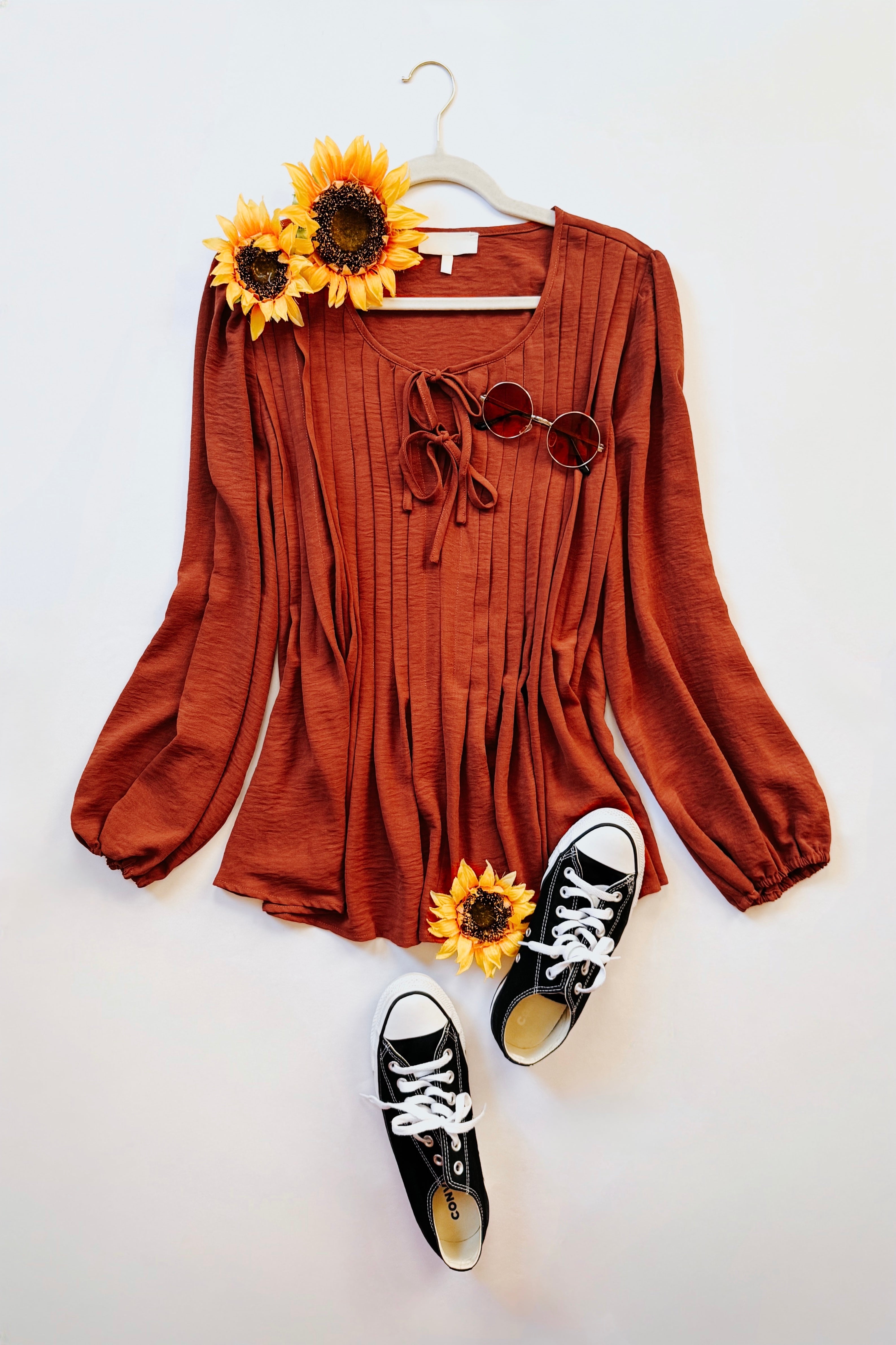 Rust-colored pleated blouse with long balloon sleeves, tie neckline, styled with sunglasses and black canvas sneakers on white background