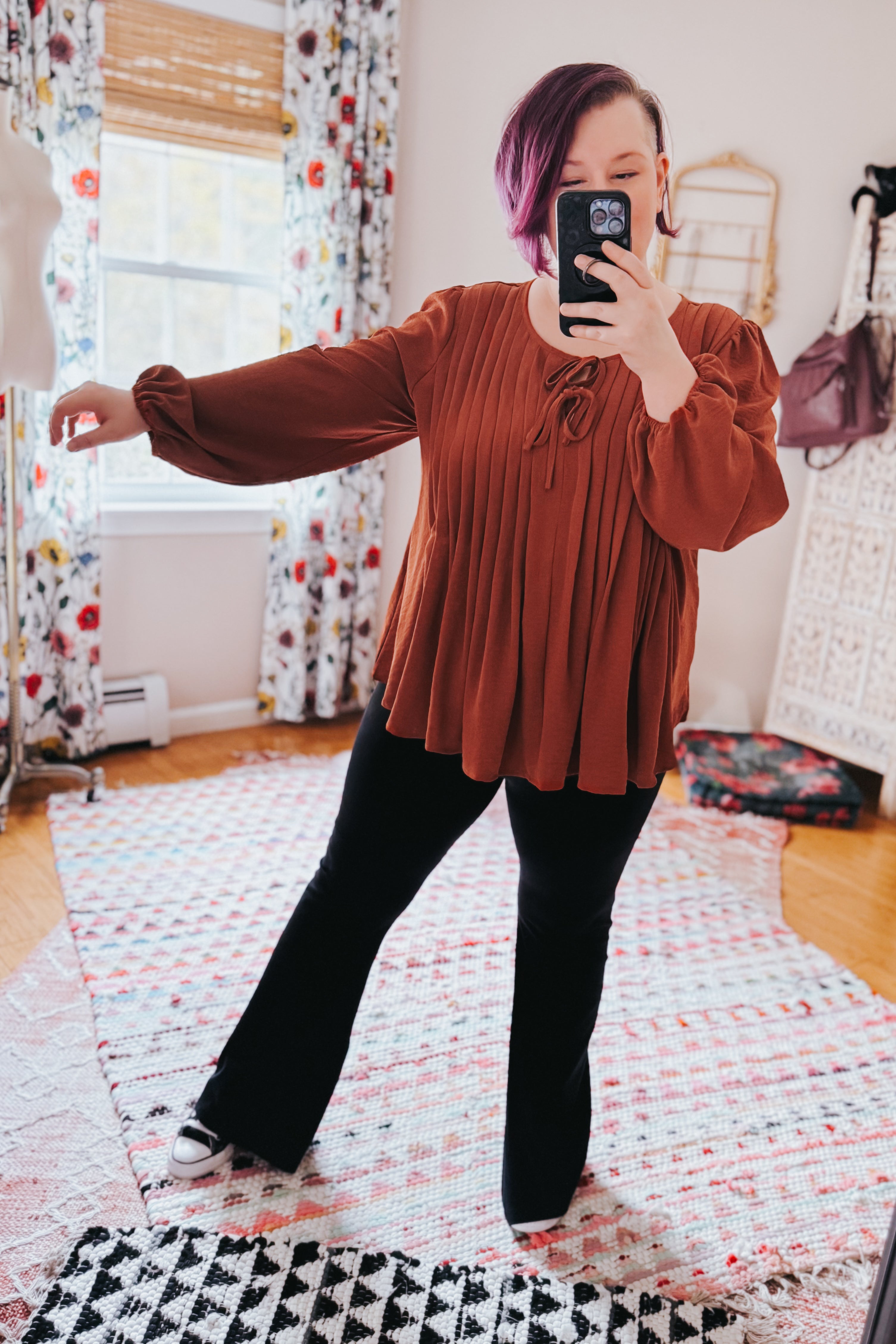 Woman wearing copper pleated blouse with long puffed sleeves and tie neckline paired with black pants standing indoors