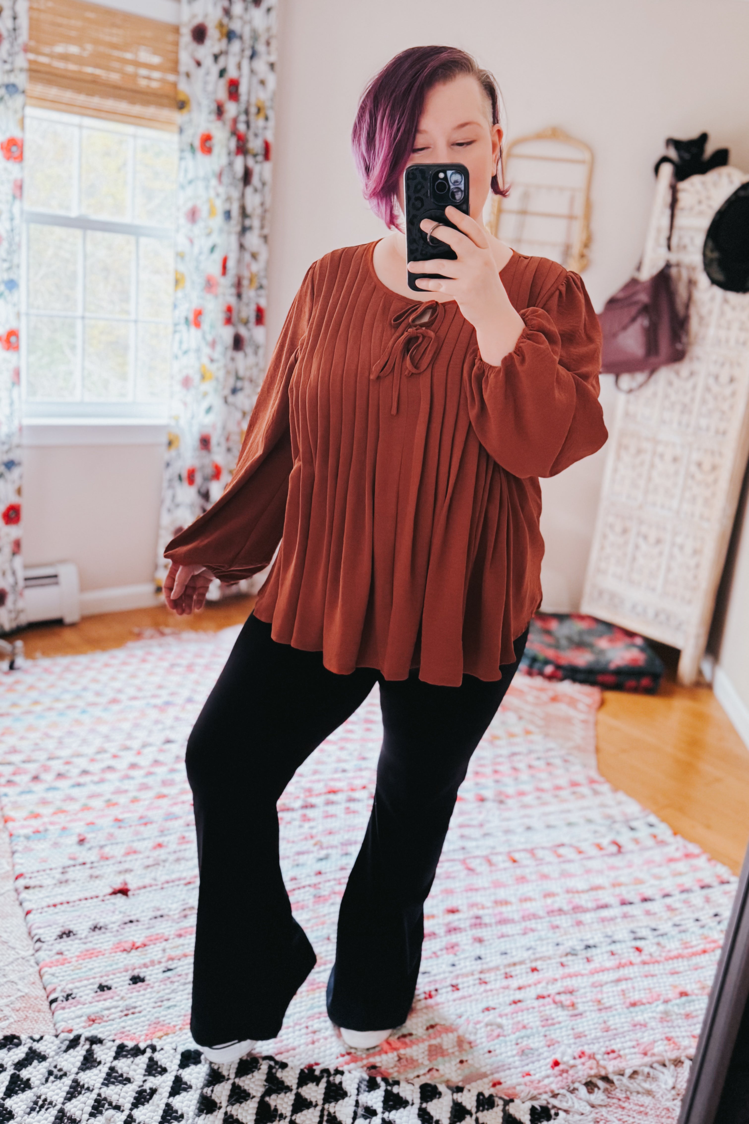 Woman taking mirror selfie indoors wearing copper pleated blouse with long sleeves and tie detail paired with black pants