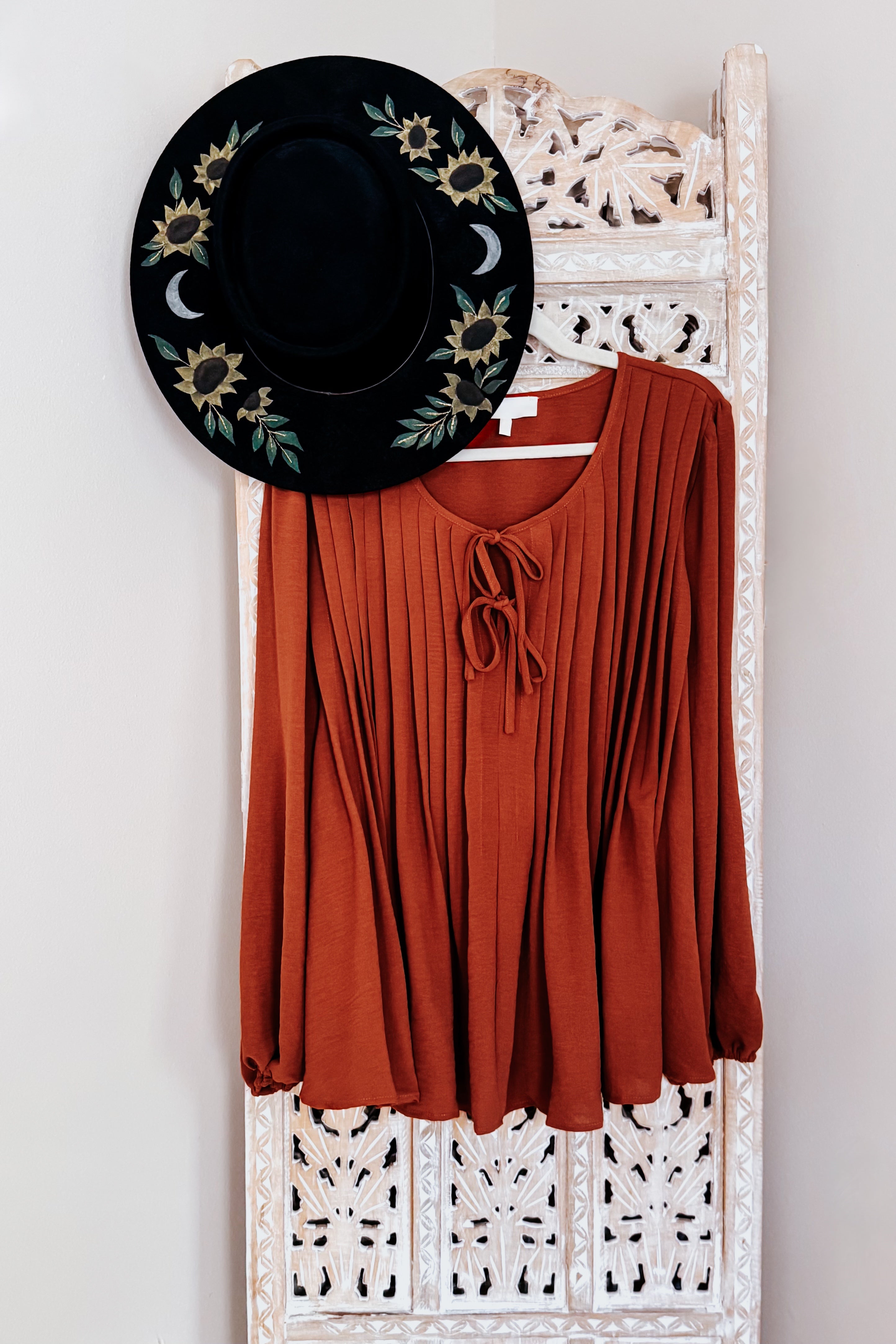 Copper-colored pleated blouse with long sleeves and keyhole tie neckline hanging on carved white wooden screen with black floral hat