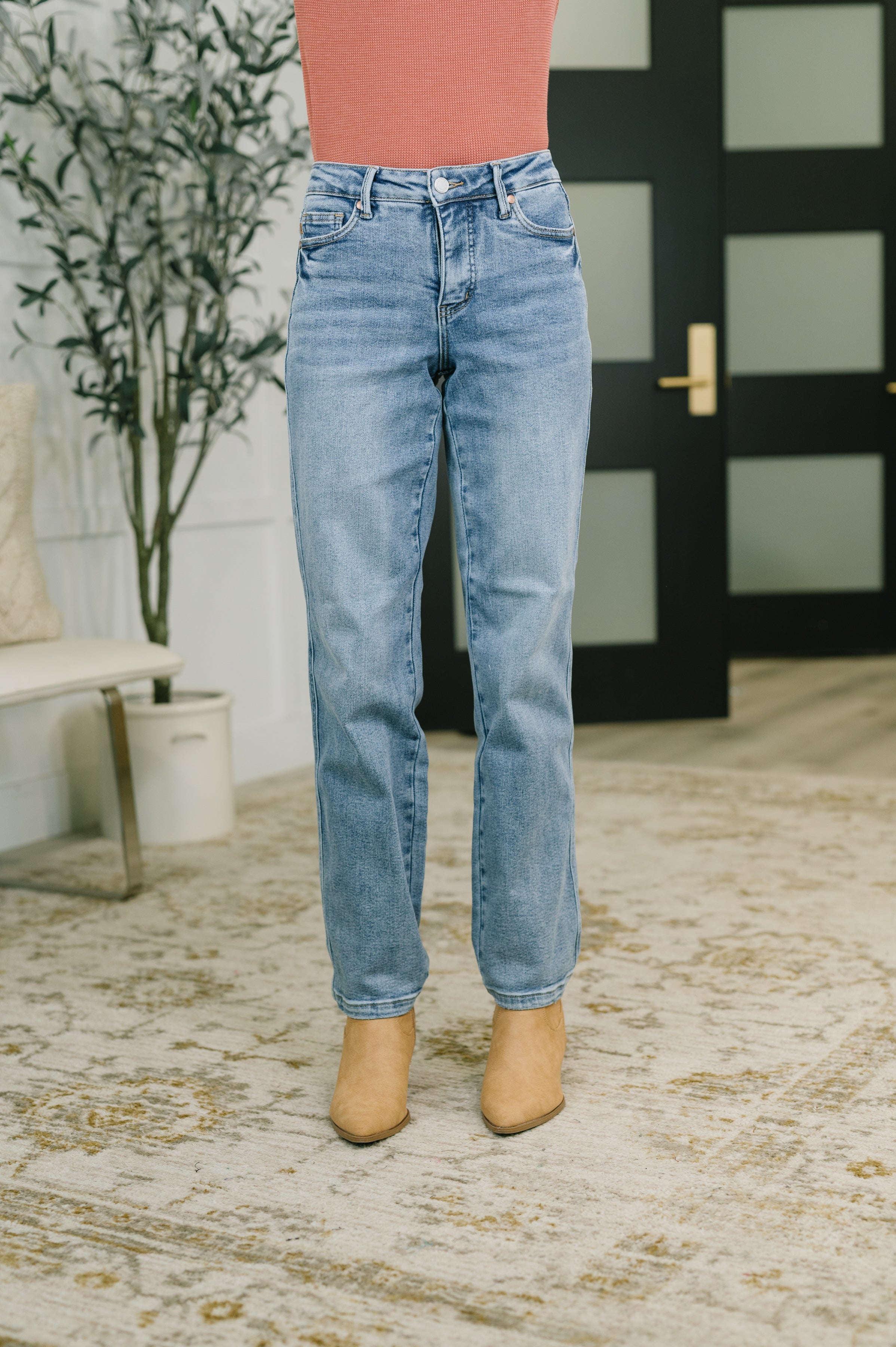 Model wearing blue mid rise straight leg jeans with a fitted waist and tan ankle boots on a patterned rug indoors