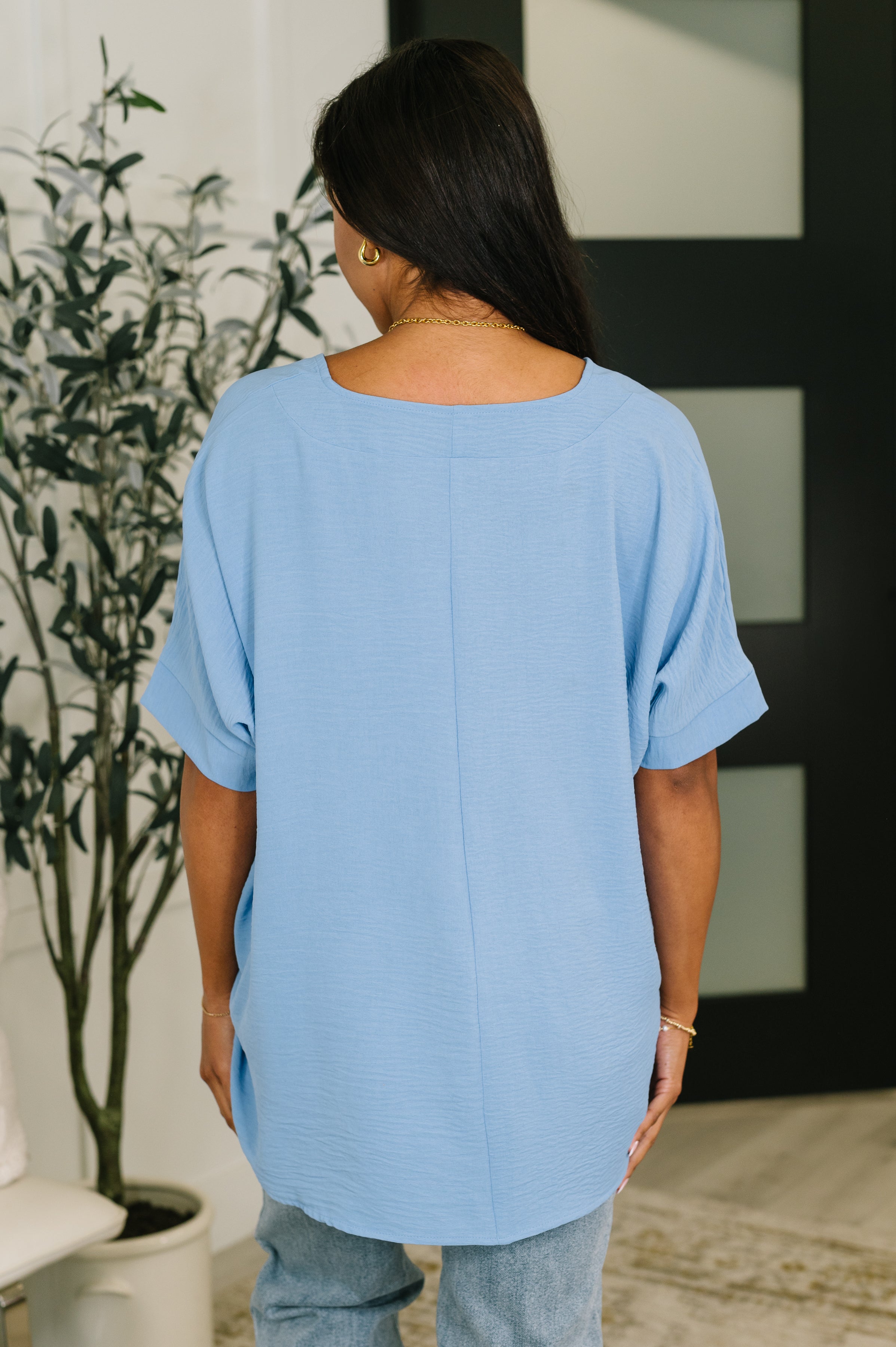Woman wearing cornflower blue crinkle woven short sleeve top with rolled cuffs standing indoors seen from behind