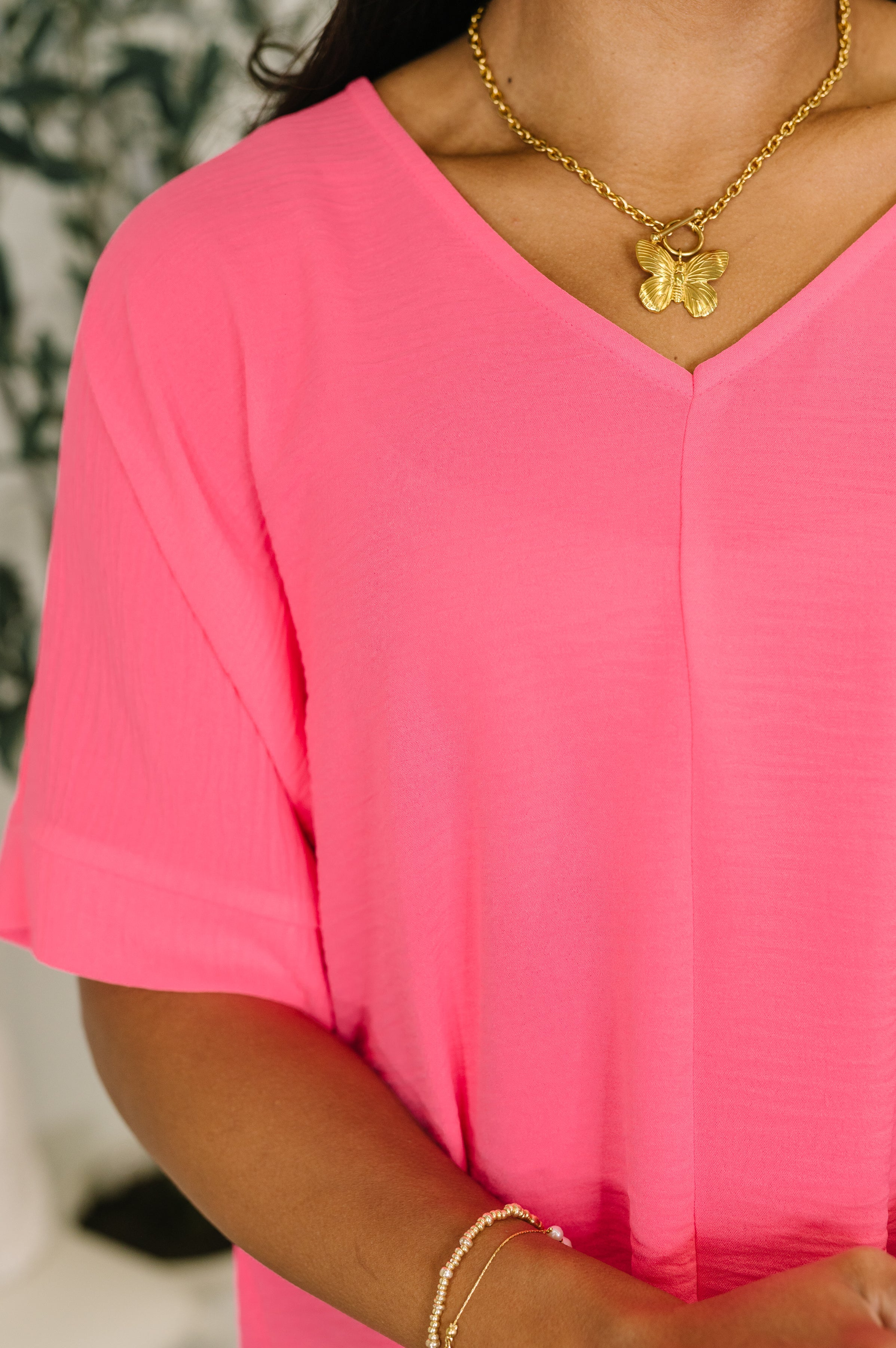 Close-up of vibrant neon fuchsia crinkle woven V-neck top with loose fit and cuffed short sleeves worn with gold butterfly necklace